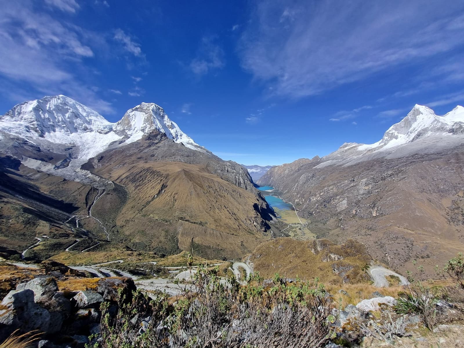 Two turquoise lakes, three snow peaks, and a road that earns every curve. Wide aerial view of the santa cruz trek route showing a winding mountain road descending past two vivid turquoise glacial lakes nestled in a high Andean valley, flanked by snow-capped peaks under a deep blue sky