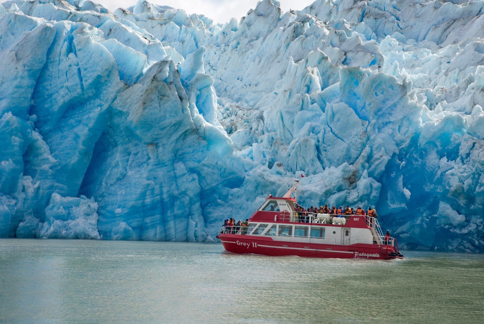 The boat is tiny. The glacier is not. The Grey II tour boat packed with passengers in orange life jackets floats directly in front of the towering blue ice face of Grey Glacier during the short w trek boat excursion on Lake Grey