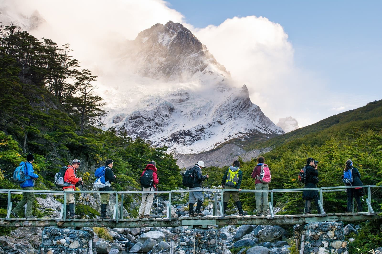 Nobody stops for long on this bridge, but nobody moves fast either. A group of hikers with backpacks and trekking poles crosses a wooden footbridge on the short W trek, with a snow-covered granite peak rising through clouds above lenga beech forest in Torres del Paine