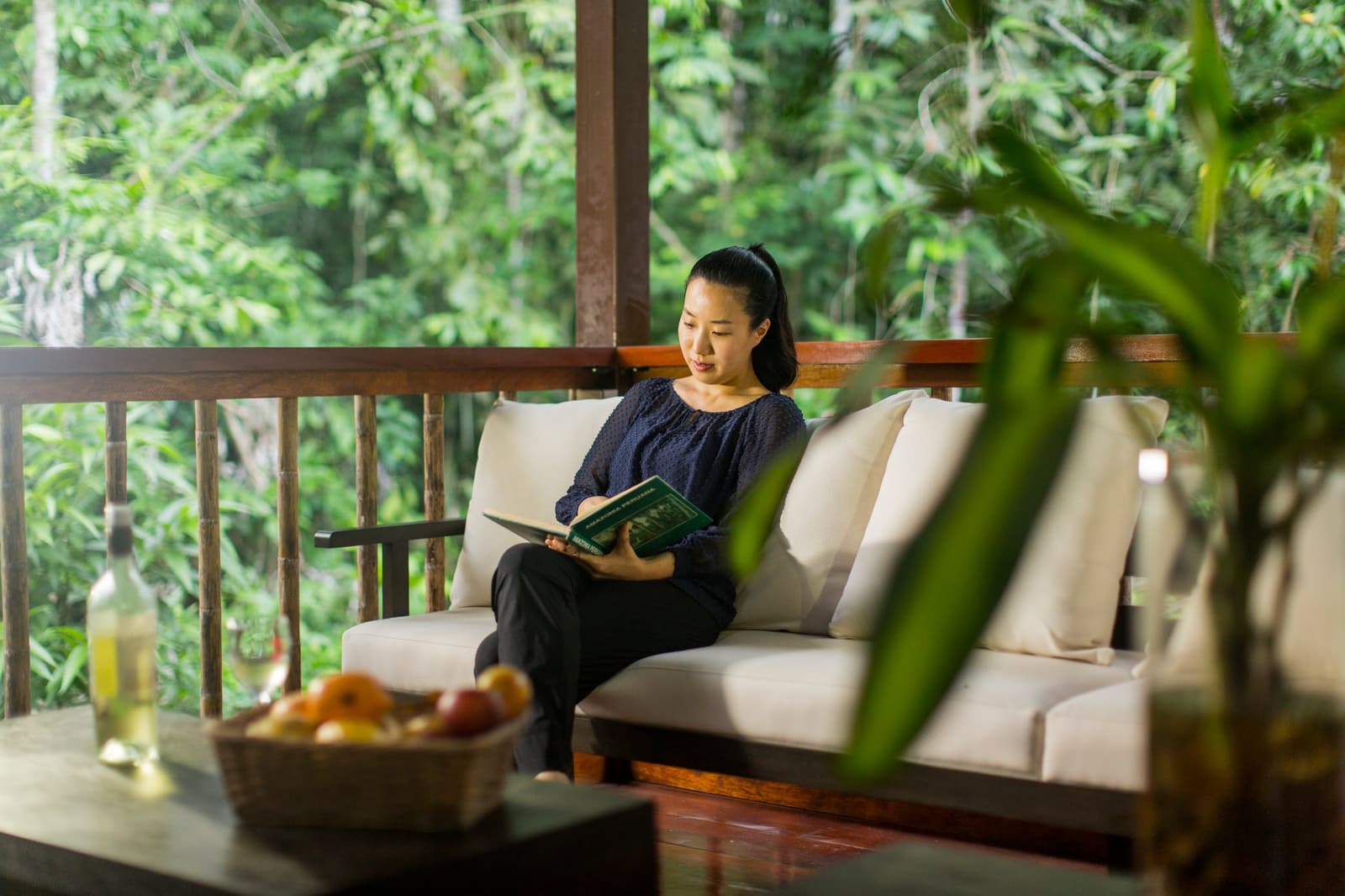 A good book, a bowl of fruit, and the Amazon just past the railing. A woman reads a book on a cushioned outdoor sofa on a screened jungle terrace at the Tambopata Research Center Peru, with dense green rainforest canopy visible behind the wooden railing, a bowl of fruit and a bottle on the coffee table in the foreground
