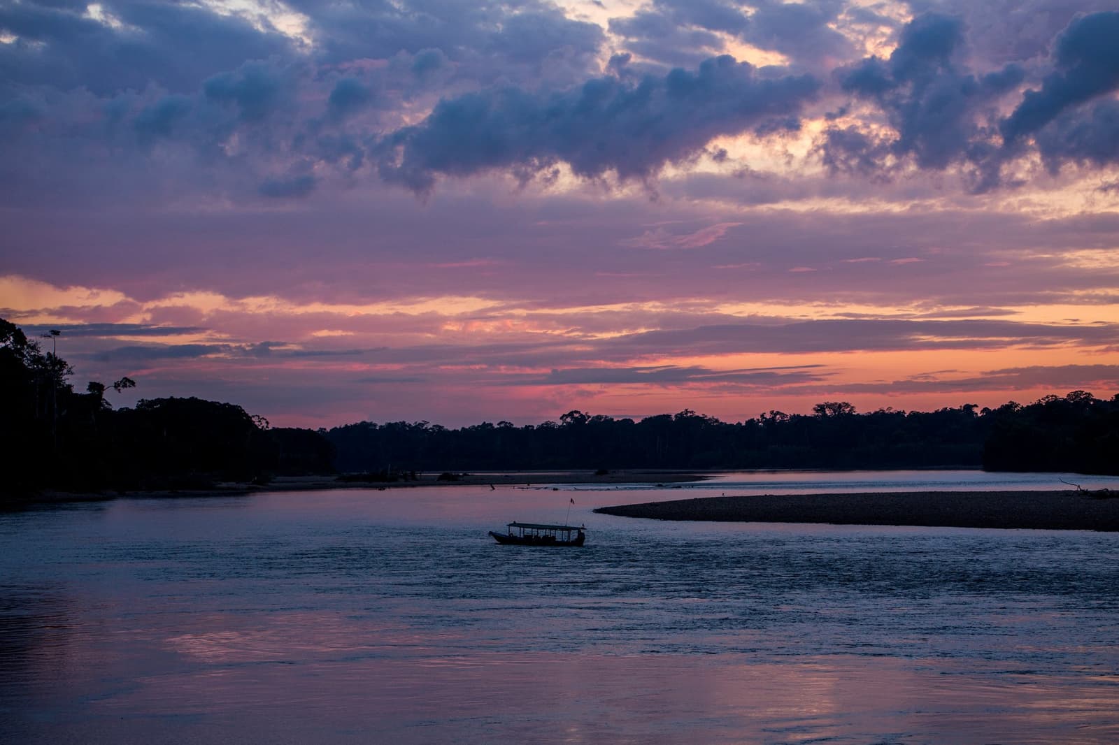 Last light, a slow current, and nowhere else to be. A silhouetted wooden boat drifts on a wide Amazonian river at dusk near the Tambopata Research Center Peru, with pink and violet clouds reflected on the calm water and a dense forest treeline along both banks