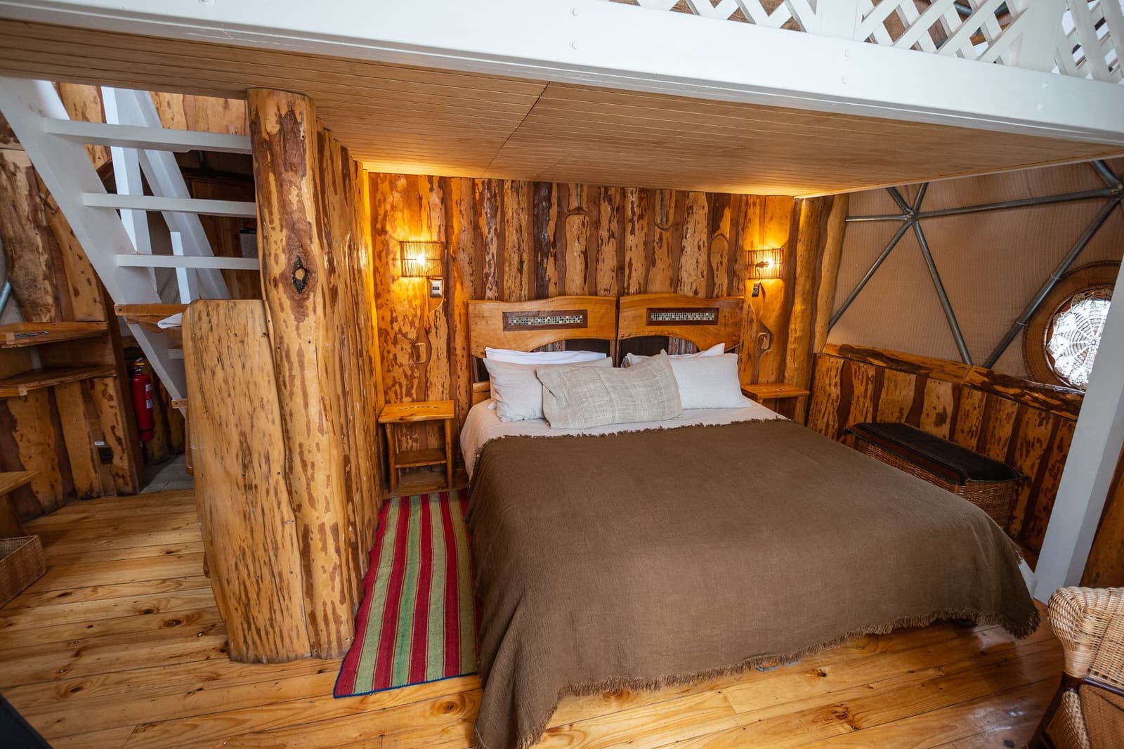 Log columns, warm light, a bed that earns its keep after a long trail day. Photo: Suite Dome Loft Interior of a geodesic dome loft bedroom on the torres del paine 4 day all inclusive tour, featuring a double bed with a brown woolen throw, rustic log columns, reclaimed-wood wall cladding, warm wall sconces, a striped rug on timber floors, and a staircase leading to an upper level