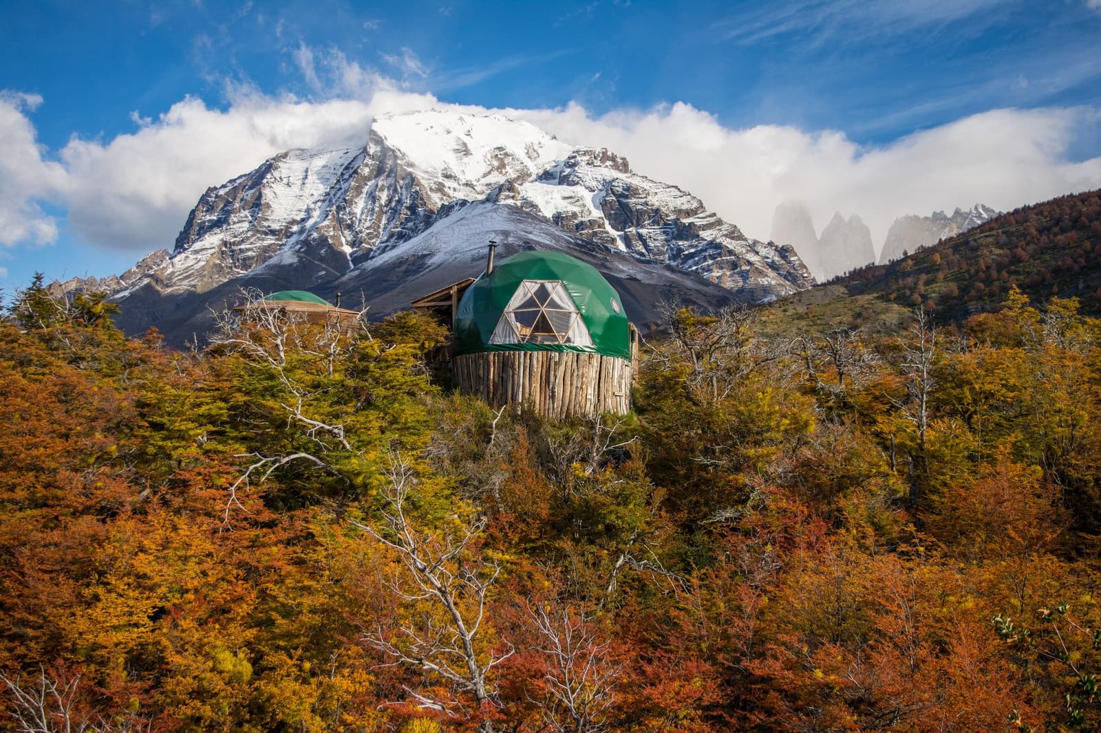 Autumn in Patagonia makes the walk to dinner feel like a reward in itself. Photo: Suite Dome A green geodesic dome on a wooden log base rises above autumn lenga forest in gold and red, with snow-capped peaks and the distinctive granite towers of Torres del Paine visible behind it — the kind of torres del paine 4 day all inclusive tour base camp that earns its view.