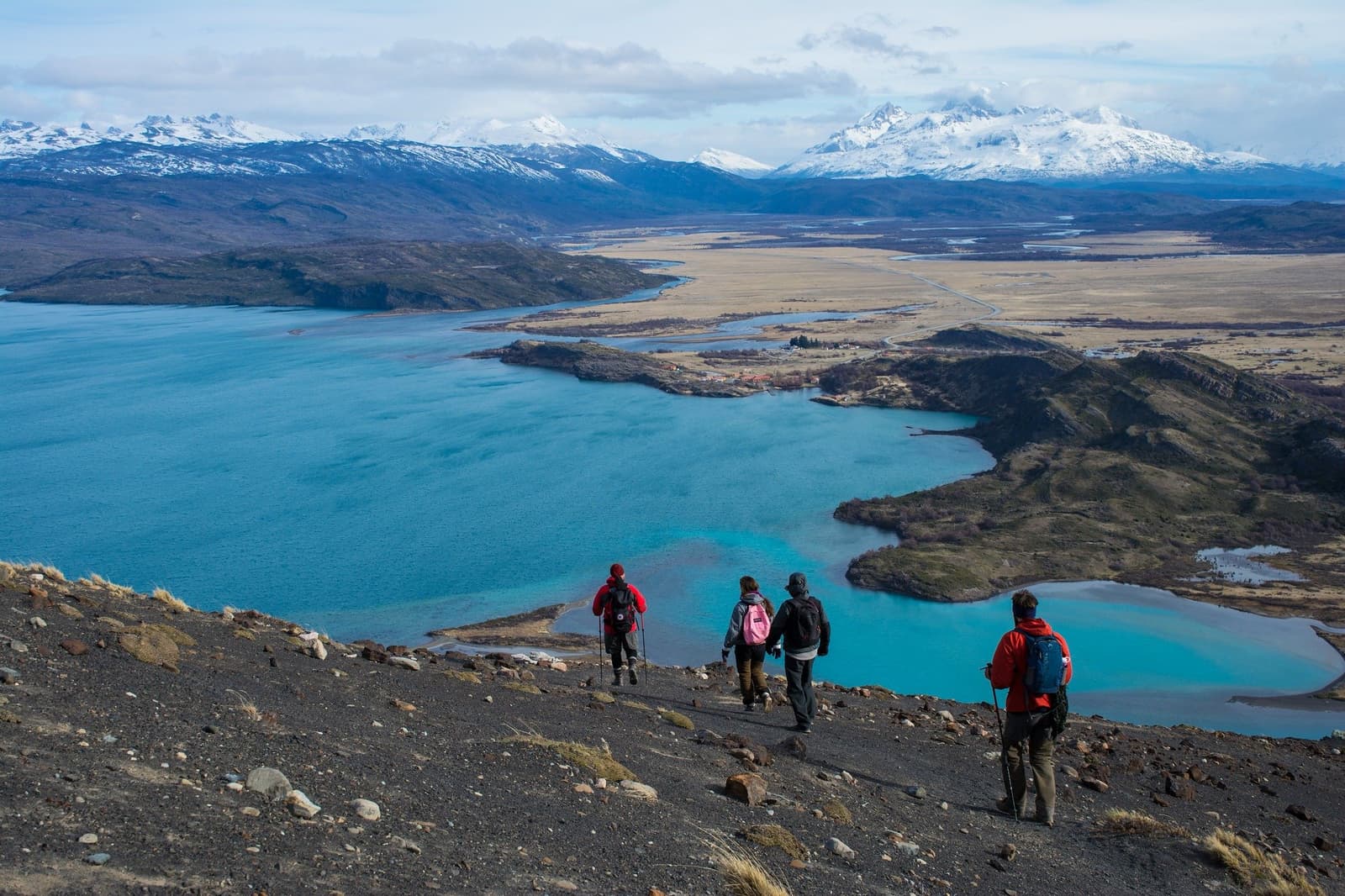 Four people, one enormous view, and a lot of descent still ahead. Four hikers with trekking poles descend a rocky volcanic slope on a torres del paine 4 day all inclusive tour, with vivid turquoise glacial lakes, golden Patagonian steppe, and snow-capped Andes peaks stretching to the horizon below them