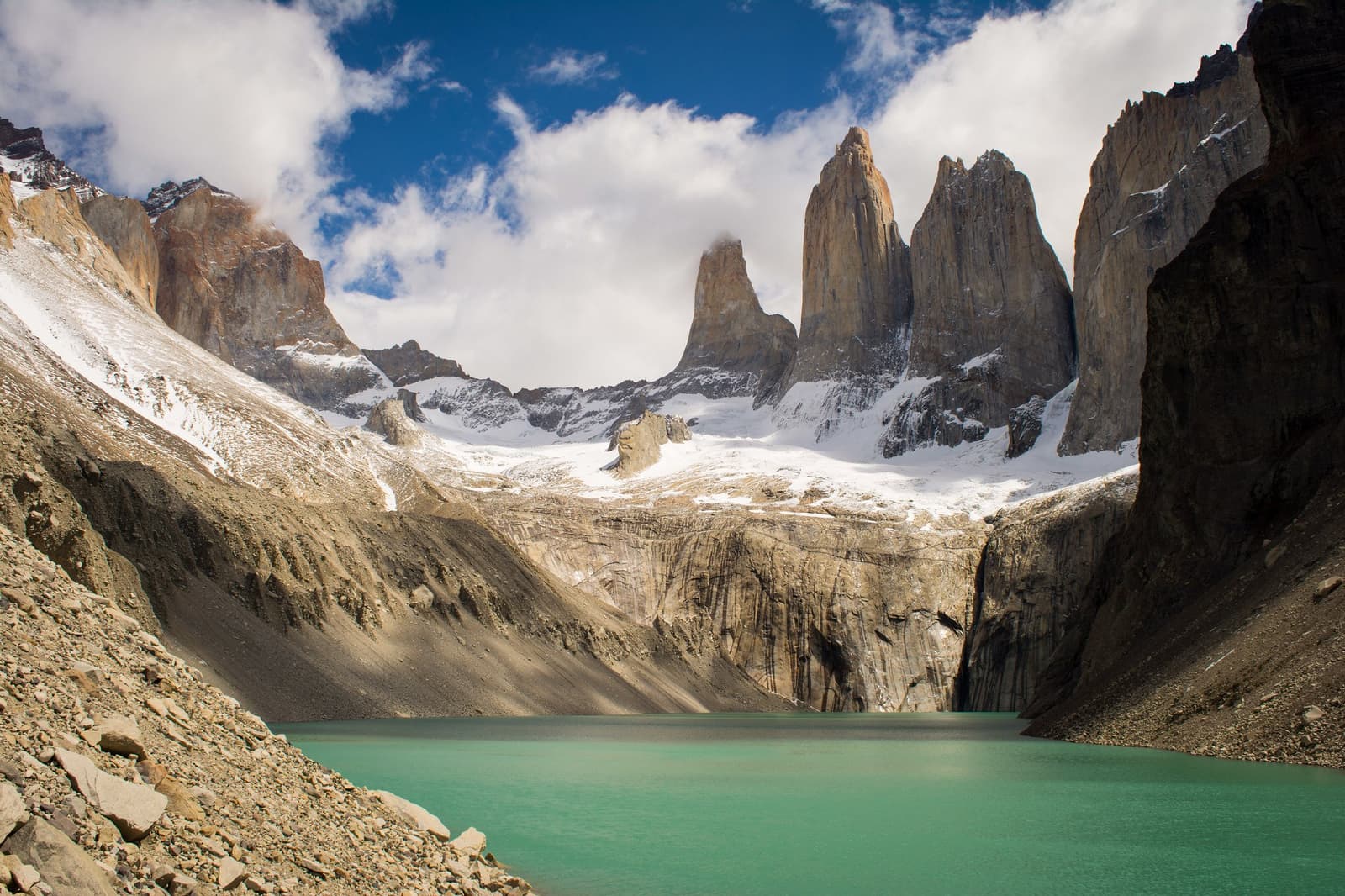 All that climbing, and then this. The lake earns its colour. Turquoise glacial lake at the base of the Torres del Paine W trek, with the three granite towers rising above snow-dusted moraines under a partly cloudy blue sky