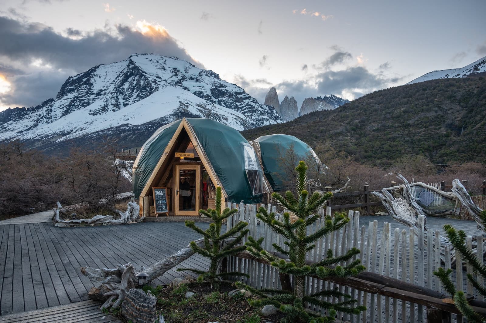 First stop, best view. The welcome dome sets the tone for everything ahead. Green geodesic welcome dome at EcoCamp Patagonia on the torres del paine w trek, with the iconic granite towers and a snow-dusted mountain visible behind it at dusk