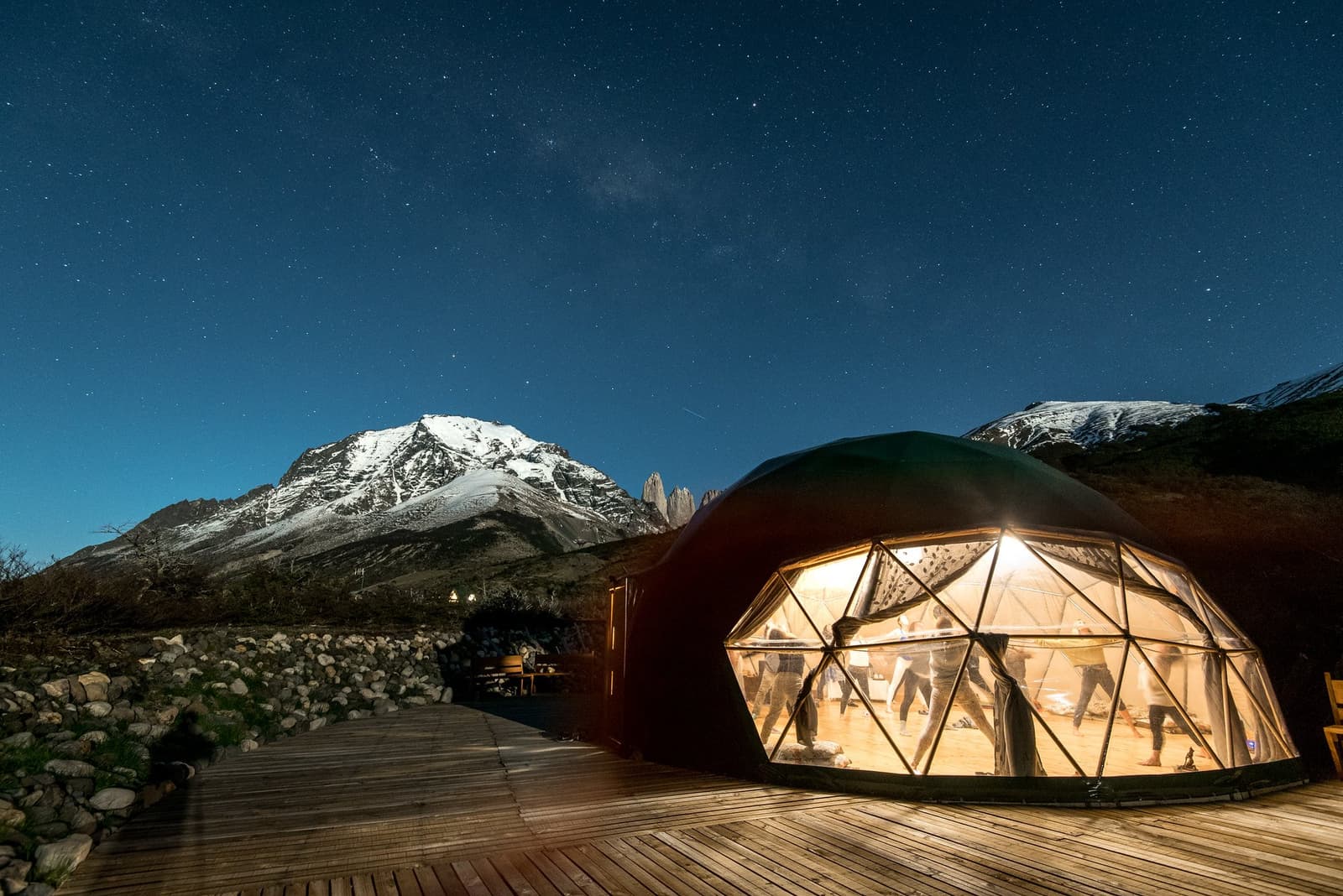 Uma aula de yoga depois da trilha, com o céu cuidando da decoração. Photo: Yoga Dome Uma cúpula geodésica de yoga iluminada à noite no Torres del Paine W Trek, com silhuetas de pessoas em movimento no interior, montanhas nevadas e um céu estrelado ao fundo