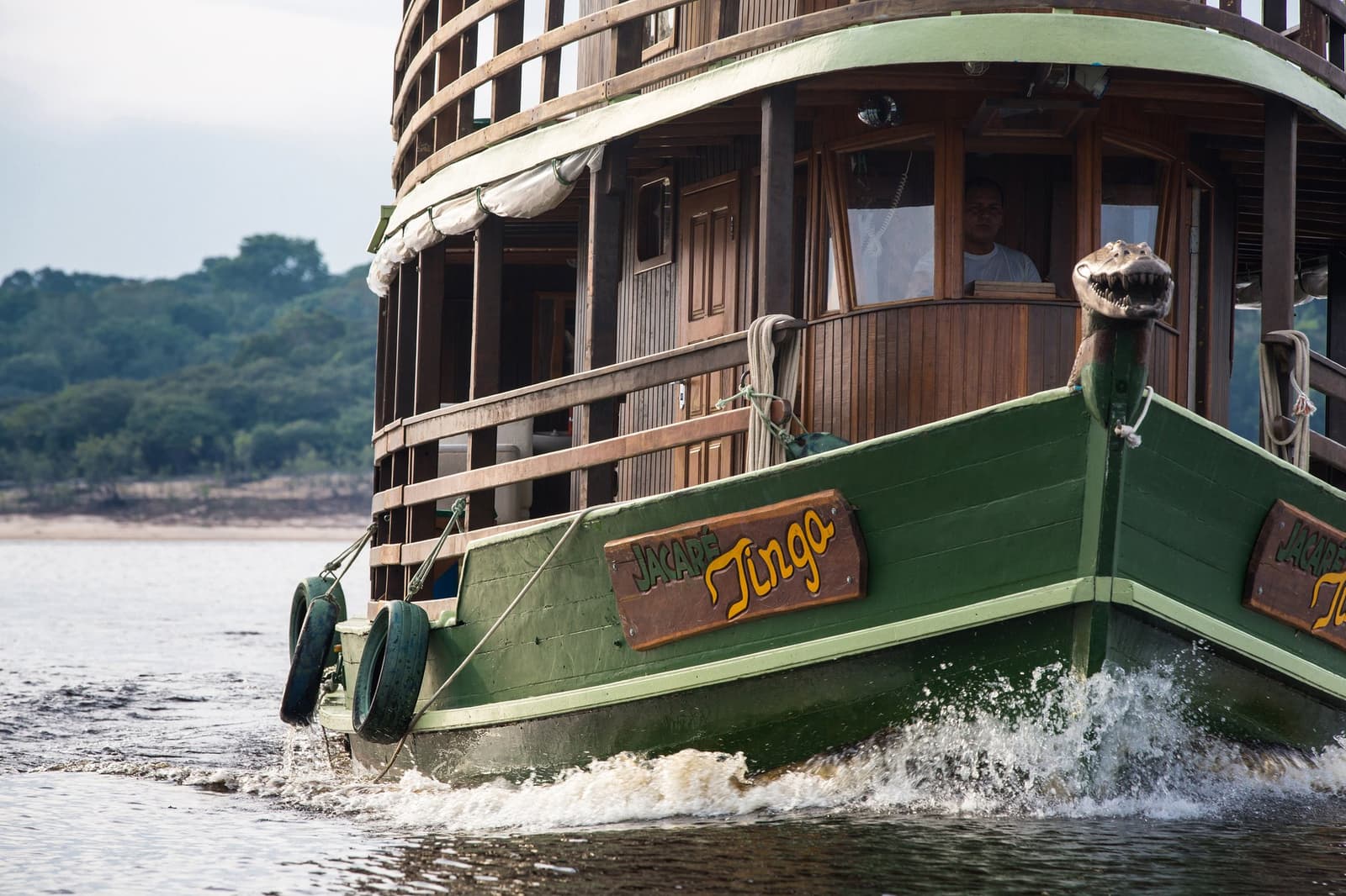 She has personality, a caiman figurehead, and a river to explore. Photo: sitah The green wooden bow of the Jacaré-Tinga boat cutting through dark river water on an amazon cruise trip in Brazil, with a caiman figurehead at the prow and forested banks in the background