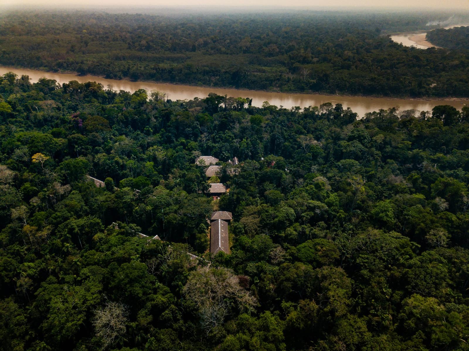 The lodge is in there, promise. The forest just makes you earn the view. Aerial drone view of the Posada Amazonas lodge buildings half-hidden beneath a dense rainforest canopy, with a wide brown river curving through the trees in the background on this amazon tour from puerto maldonado