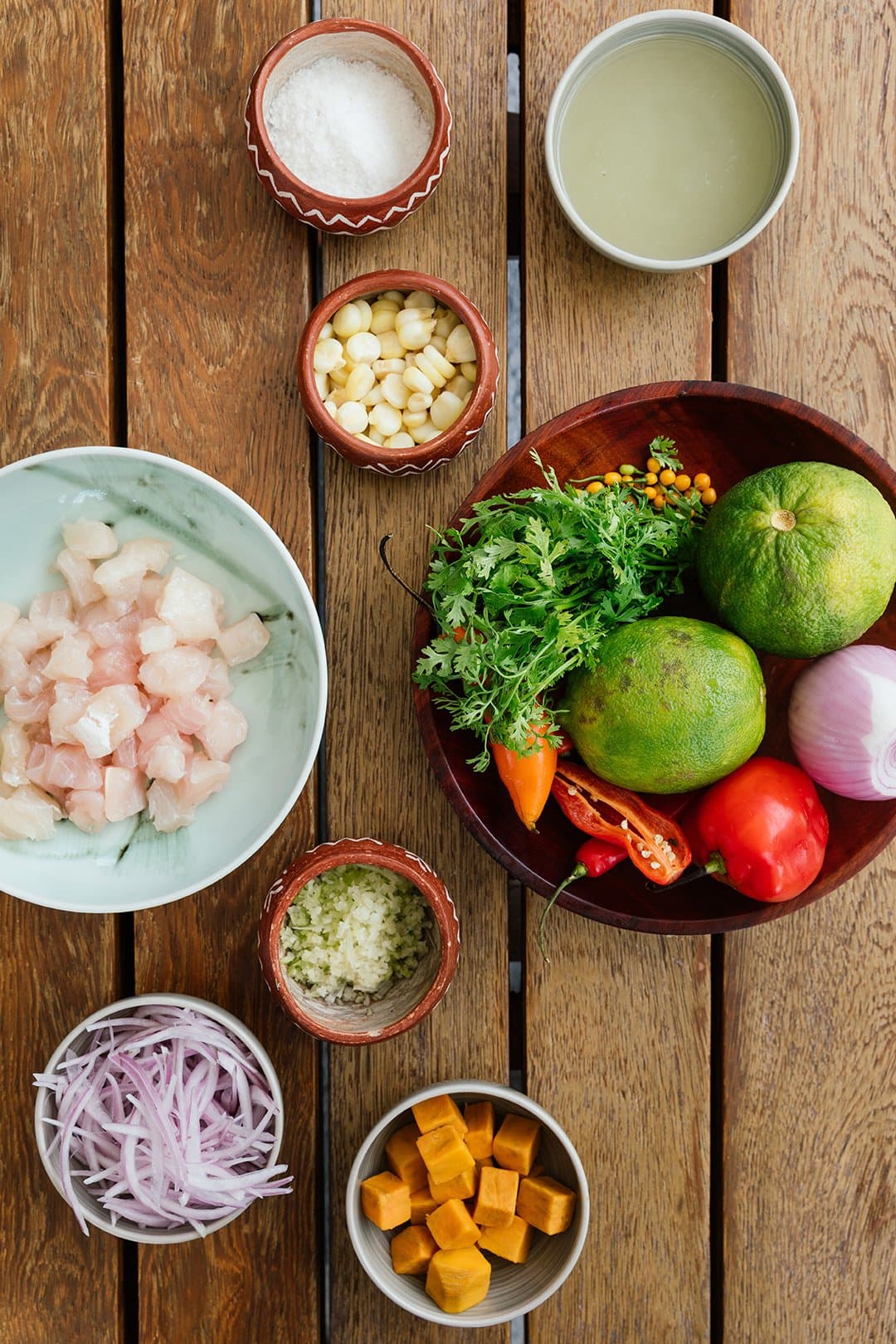 Every bowl on this table has a story from the market that morning. Photo: Aqua Expeditions Flat-lay of fresh ceviche ingredients on a wooden table for the aria amazon cruise kitchen: raw white fish cubes, sliced red onion, chopped garlic, corn kernels, diced orange sweet potato, halved chili peppers, tomato, limes, fresh cilantro, small berries, salt, and a bowl of citrus juice