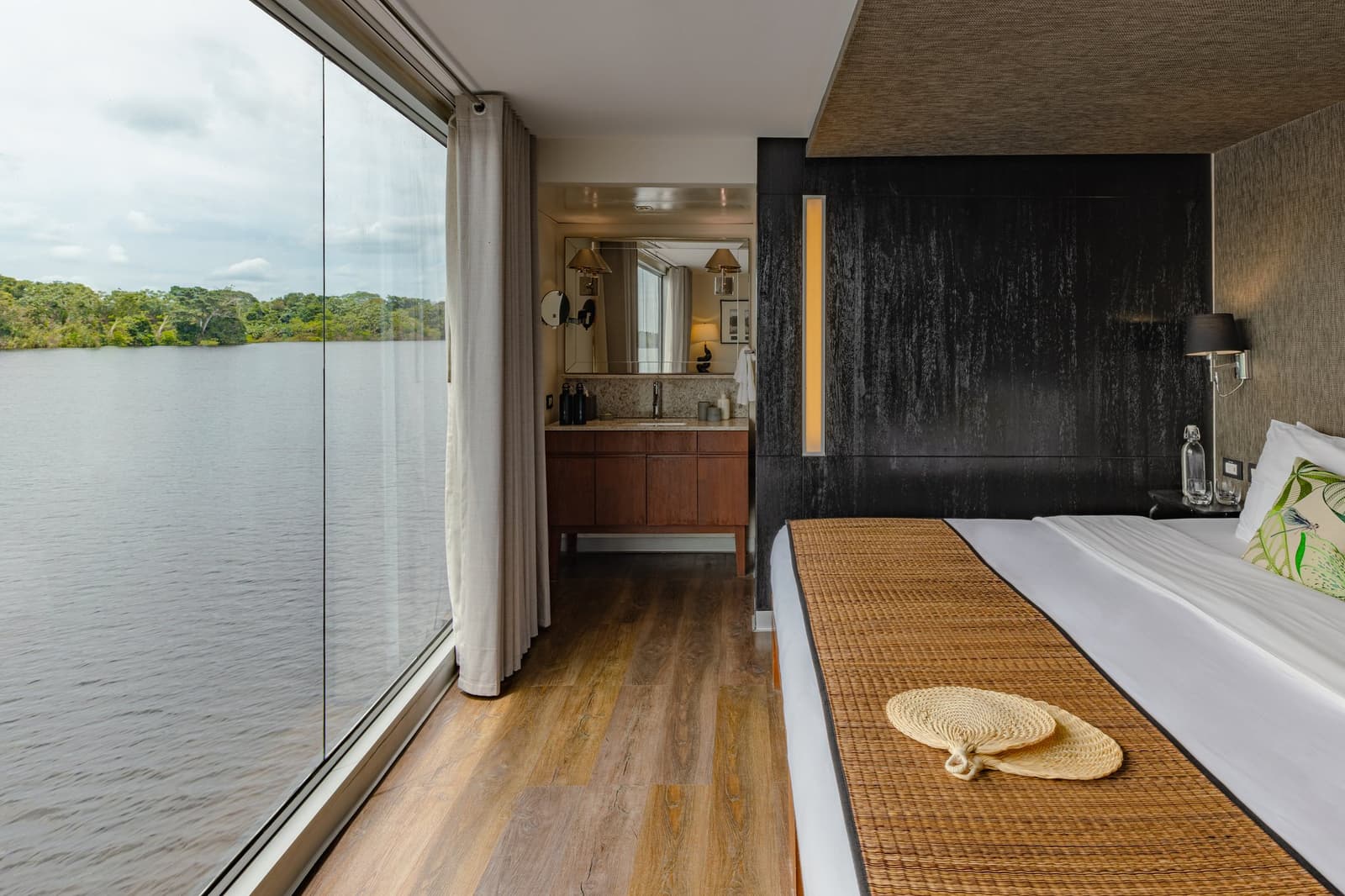 Your bedroom, the river, and not a single reason to rush. Photo: Francois Moreau Elegantly appointed suite cabin on the Aria Amazon cruise, with a king bed, woven runner, straw fan, dark textured accent wall, wood vanity visible through the open bathroom door, and floor-to-ceiling glass windows overlooking a calm river and dense jungle canopy