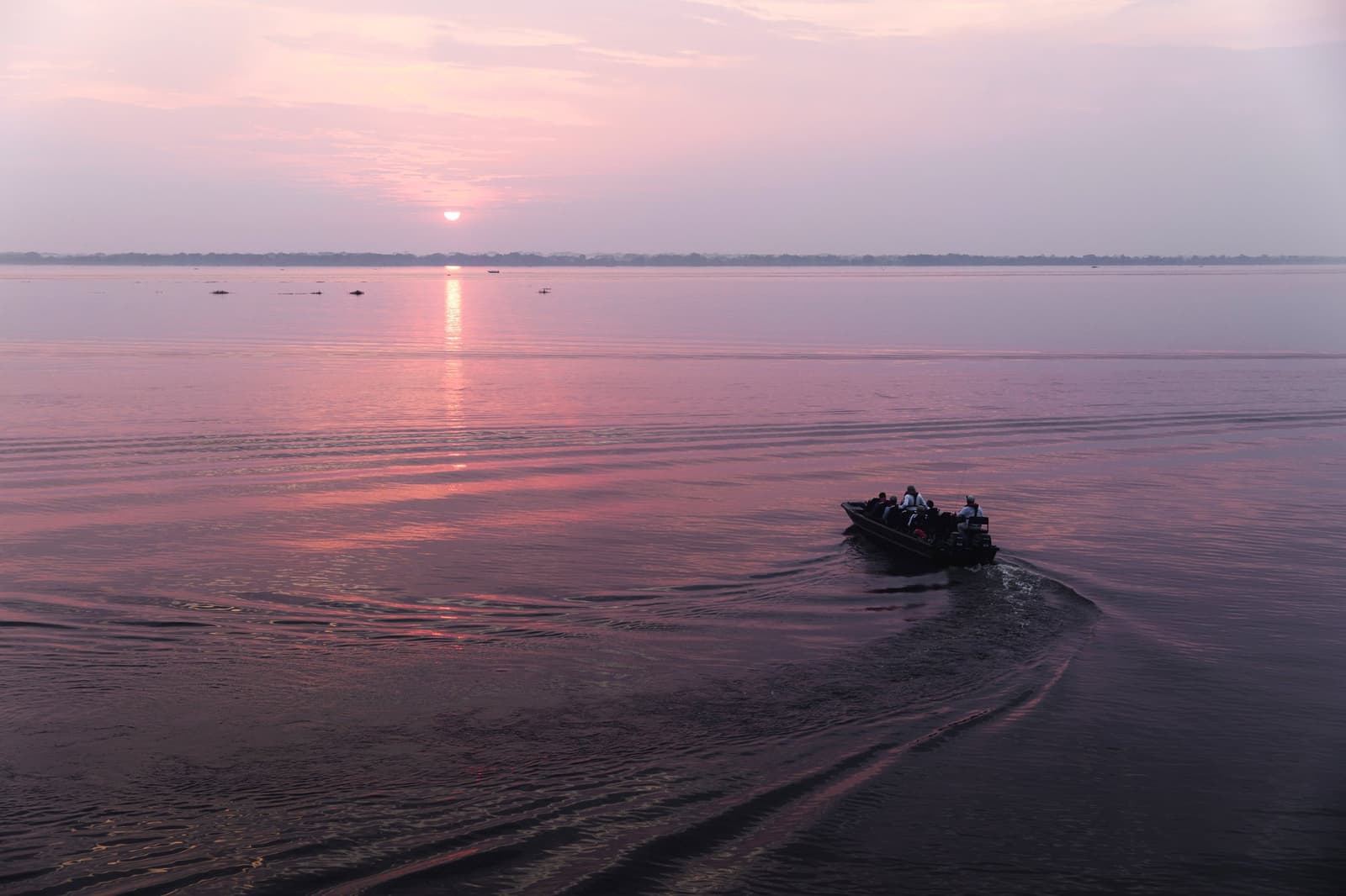 Pink light, calm water, and somewhere out there, a dolphin fin breaks the surface. Photo: Anthony K Do A small skiff carrying several passengers crosses a wide river at sunset during an Aria Amazon cruise, with a rose-pink sky reflected on the glassy water and a distant forested horizon
