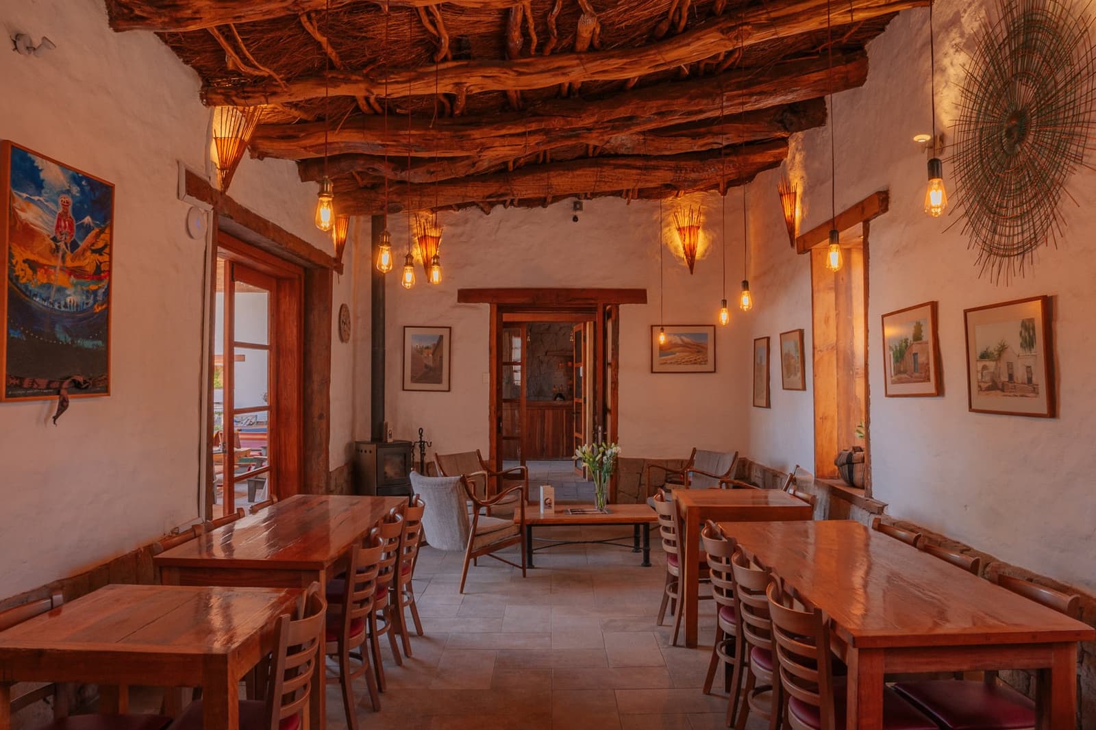 Después de dormir bajo las estrellas, esta sala se siente como llegar a casa. Comedor de adobe con techo expuesto de vigas y cañas, colgantes de bombillas Edison, mesas de madera pulida, pinturas locales en paredes encaladas y una estufa de leña