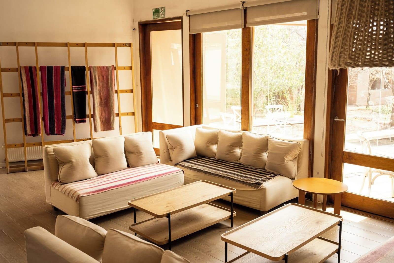 Colorful woven blankets on the rack, a deep sofa, afternoon sun. Rest earns its place on a trip like this. Sun-filled lounge area of an atacama desert tour package lodge, featuring a cream sectional sofa with striped woven blankets, wooden coffee tables, and floor-to-ceiling windows overlooking a garden courtyard