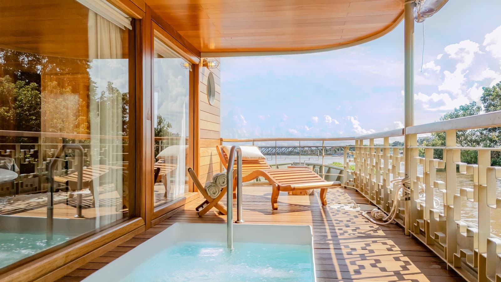 Jacuzzi, espreguiçadeira, o rio passando devagar. Algumas escolhas não precisam de ajuda. Varanda privativa do cruzeiro Delfin Amazon com banheira de hidromassagem, espreguiçadeira de teca e vista aberta para o rio sob um teto de madeira curvo
