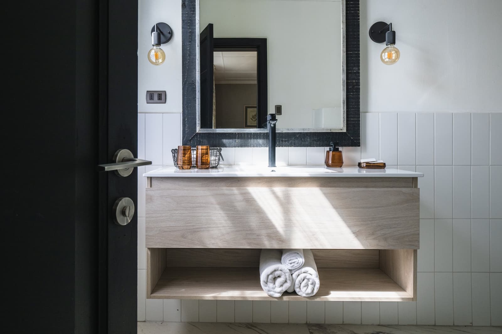 Clean lines, warm light, rolled towels. Ranch life has come a long way. Photo: CARLOS DIAZ Floating wood vanity with white countertop, black-framed mirror, amber glass toiletry accessories, rolled white towels, and Edison bulb wall sconces in a private bathroom at Estancia Cerro Guido