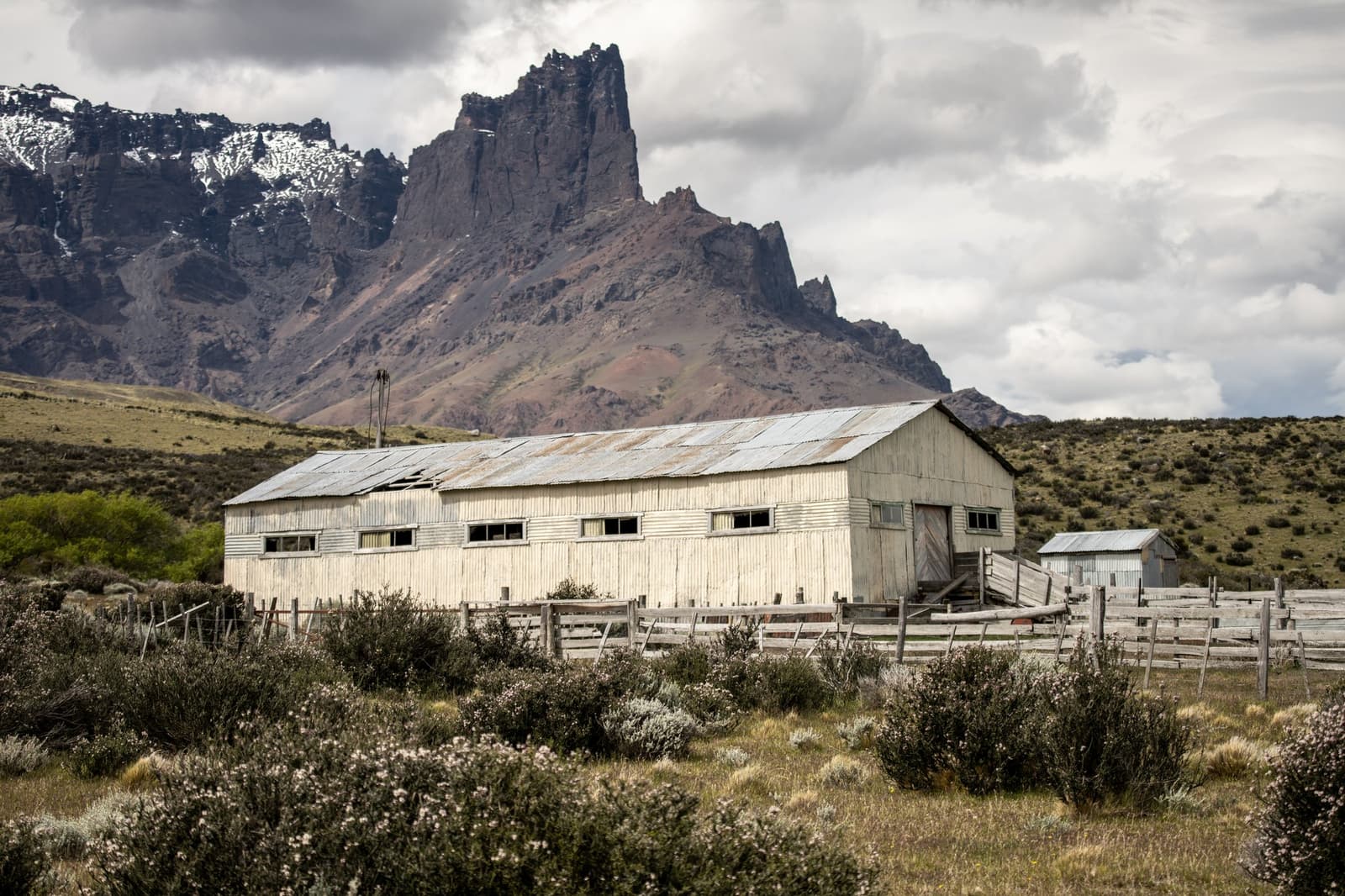 A century of wool, wind, and hard work still standing in the steppe. A weathered white corrugated-iron shearing shed with wooden livestock pens at estancia cerro guido, set against jagged Patagonian peaks and a cloudy sky