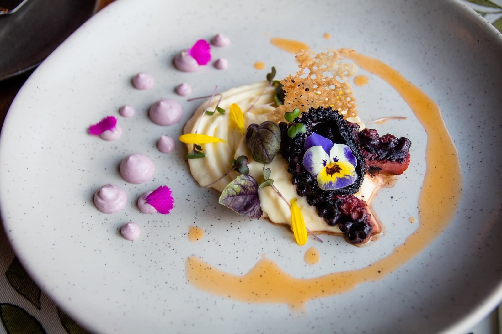 Petals, pearls, and a sauce swirl: the kitchen is clearly having fun. Close-up of a fine-dining dessert plate at Estancia Cerro Guido, featuring a cream quenelle, dark berry compote, caramelised tuile, lavender cream dots, magenta petals, yellow flower petals, micro herbs, and a fresh pansy blossom on a speckled white ceramic plate with an amber sauce swirl