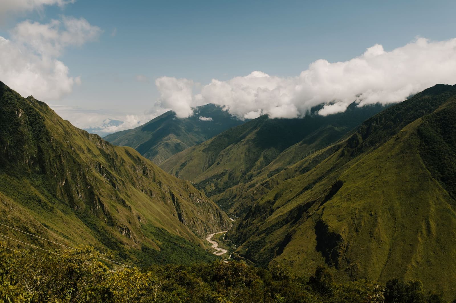 Somewhere between the rainforest and the high pass, the road hands you this. A deep green Andean valley with a winding river at its floor viewed from a high ridge on the machu picchu amazon tour, with layered cloud-forest peaks and low clouds in the distance