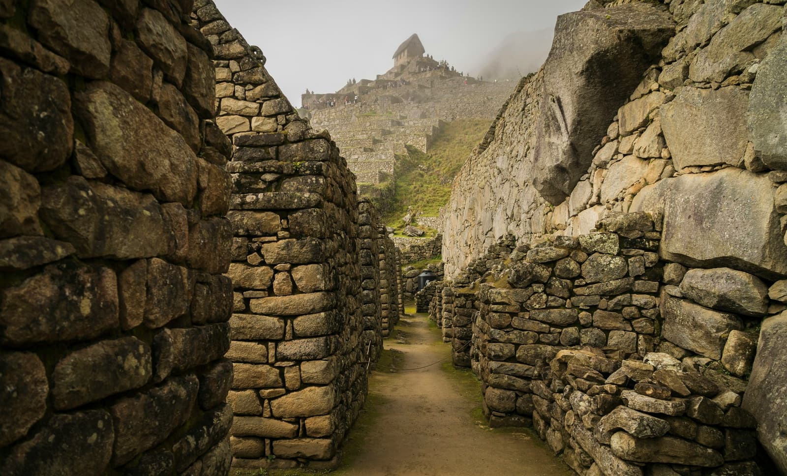 Follow any passage long enough and the citadel reveals itself, a little at a time. A narrow path runs between tall Inca stone walls inside Machu Picchu, with terraced ruins and a mist-shrouded structure visible on the hilltop above, on a machu picchu amazon tour machu picchu and amazon tours itinerary