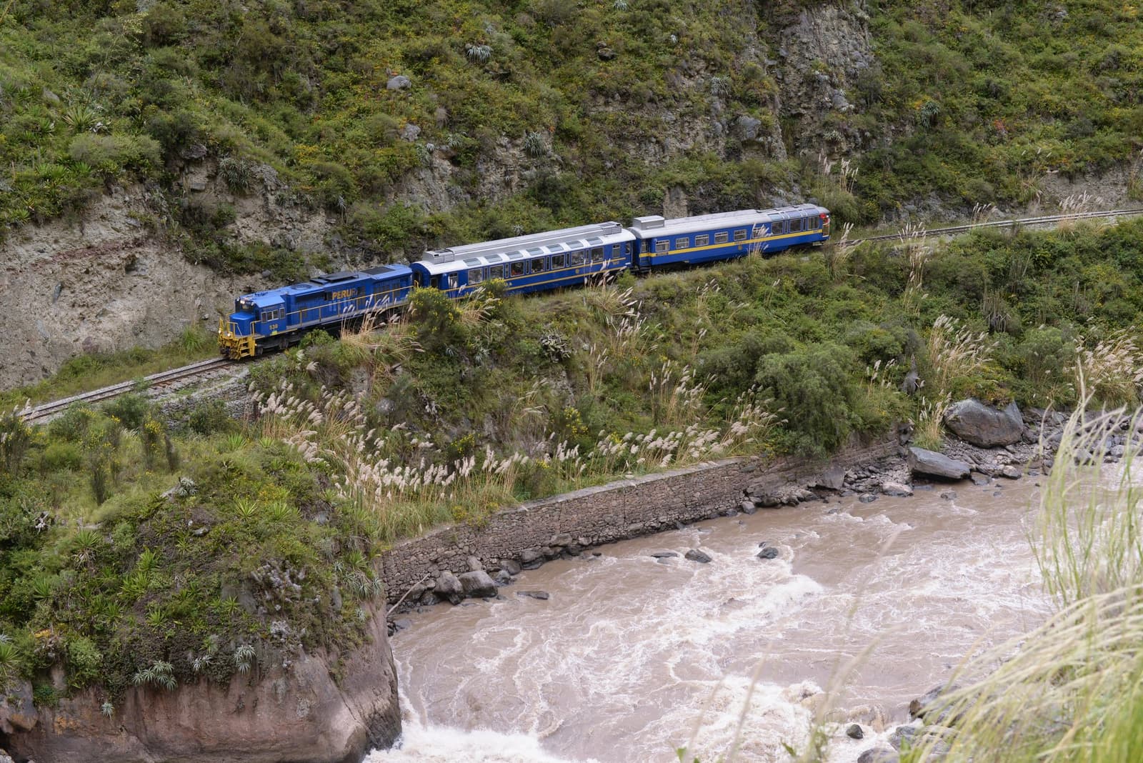 Squeezed between cliff and river, the train earns every kilometre. A blue Peru Rail train with panoramic carriages travels along a narrow ledge above the rushing Urubamba River, flanked by steep green canyon walls, on the machu picchu amazon tour machu picchu and amazon tours route through the Sacred Valley.