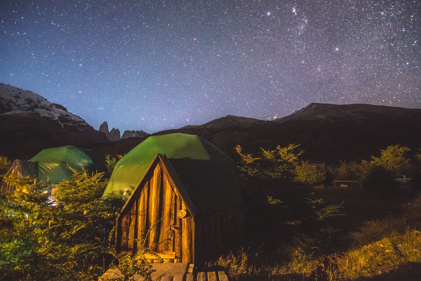 Thousands of stars overhead, and a warm dome waiting inside. Not a bad trade-off. Photo: Standard Dome Green geodesic dome tents lit from within at EcoCamp Patagonia at night, with a wooden arched doorway in the foreground, dark mountain silhouettes behind, and a dense field of stars filling the deep blue sky above the O trek in Patagonia