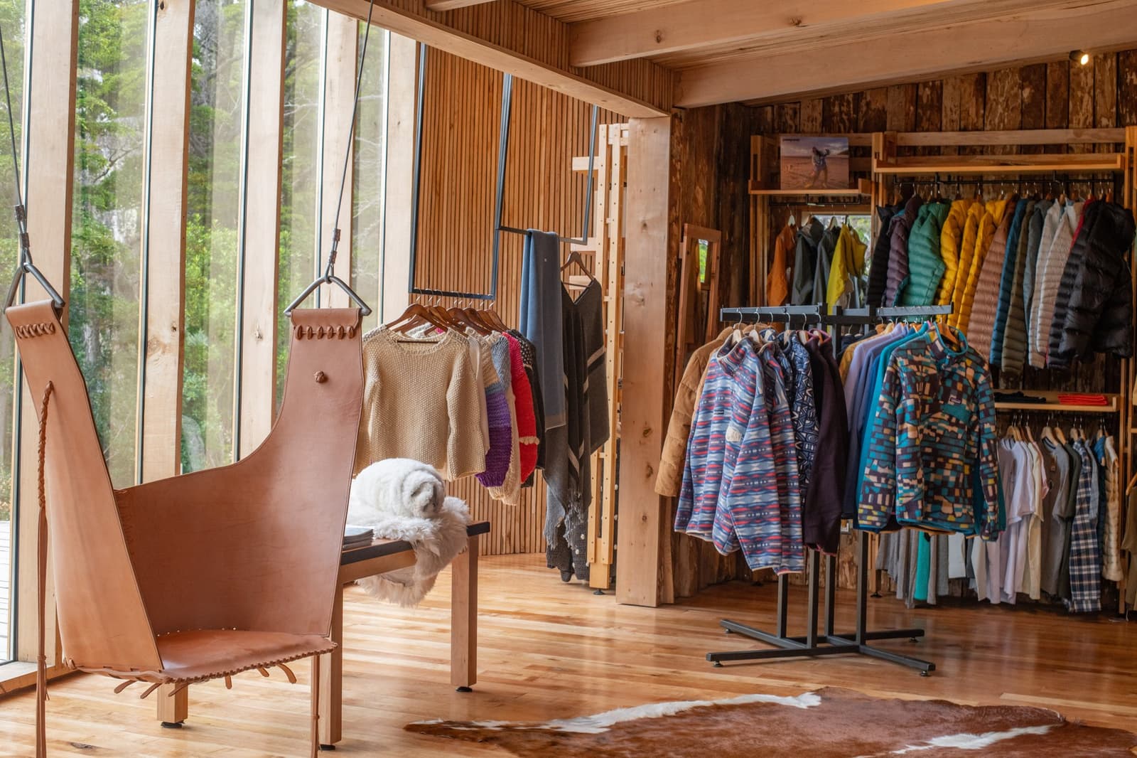 Forgot a layer? Honestly, the shop makes it easy to be glad you did. Photo: Patagonia Camp Interior of the Patagonia Camp boutique shop featuring racks of colorful fleece jackets, quilted down coats, and hand-knit wool sweaters displayed on wooden shelving, with a handcrafted leather chair and cowhide rug on a warm hardwood floor, forest visible through floor-to-ceiling windows
