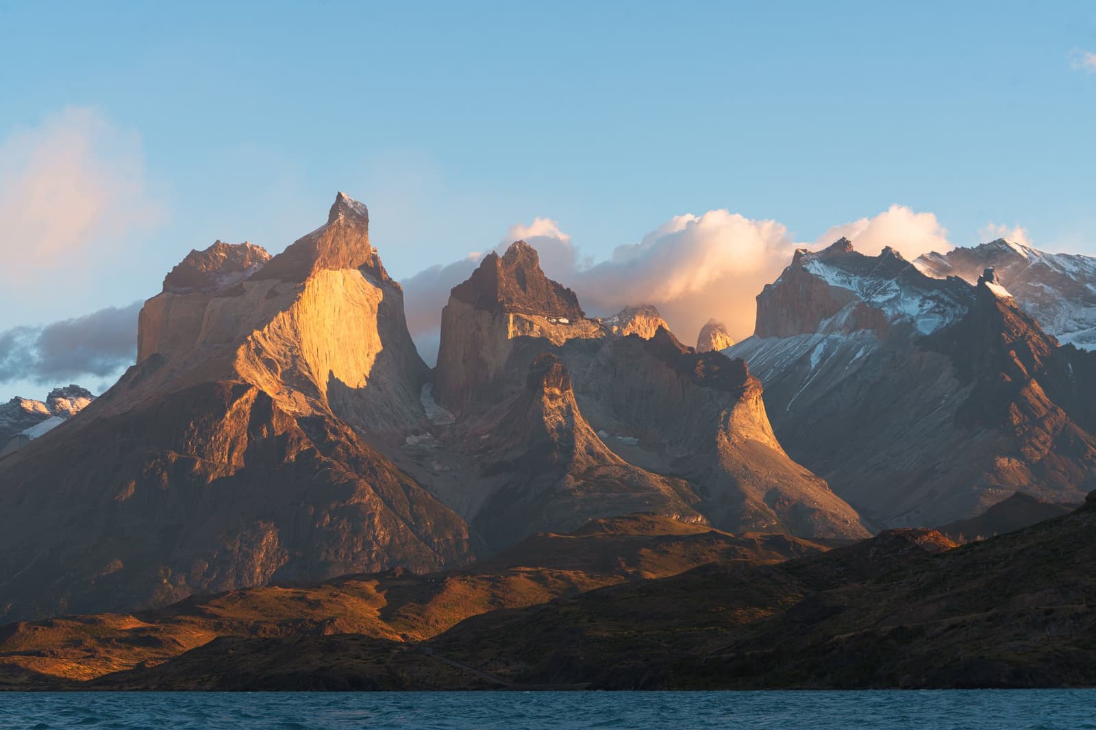 Early light picks a favourite, and today it chose the Cuernos. Photo: Patagonia Camp The Cuernos del Paine rock towers bathed in warm golden light at sunrise, viewed from the shores of Lake Toro at Patagonia Camp, with snow-dusted peaks and billowing clouds behind