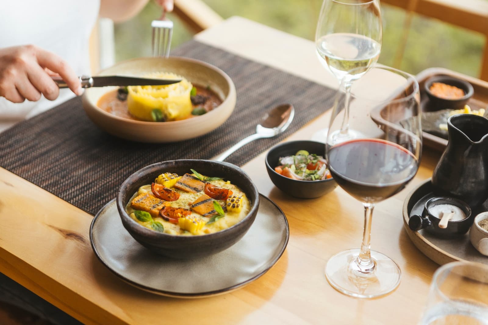 Dinner here earns its own spot on the itinerary. Photo: Patagonia Camp A plated dinner at Patagonia Camp featuring a creamy polenta bowl topped with grilled salmon, cherry tomatoes, and fresh herbs, alongside a rich meat stew with moulded rice, red and white wine glasses, and small side dishes arranged on a light wood table