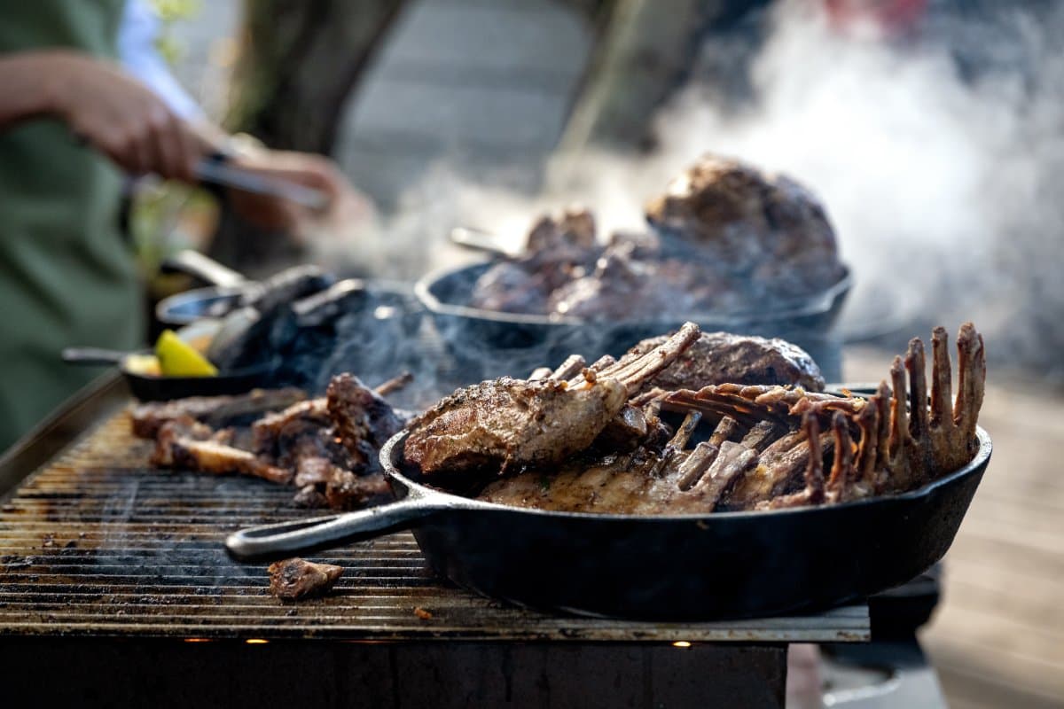 Smoke, fire, and a rack of ribs that earns its keep after a long day on the trail. Photo: Patagonia Camp Smoking cast iron skillets loaded with grilled ribs and roasted meats on an open charcoal grill at Patagonia Camp, with a cook working in the background