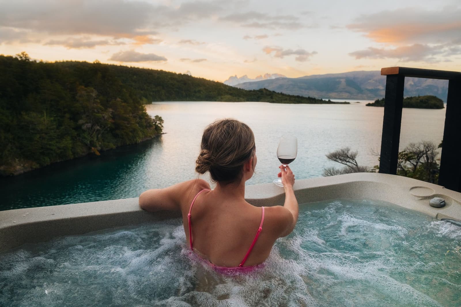 Red wine, warm water, cold air outside. Some afternoons earn this. Photo: Patagonia Camp A woman soaks in an outdoor jacuzzi at Patagonia Camp, holding a glass of red wine while looking out over turquoise Lake Toro toward distant snow-capped peaks at sunset