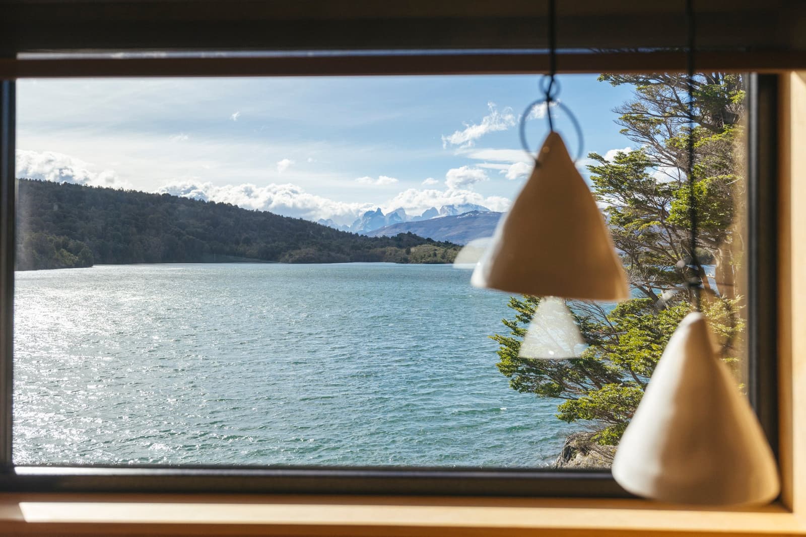 Turns out the best seat in the house has no seat at all. Photo: Patagonia Camp View through a wooden-framed cabin window at Patagonia Camp, showing the turquoise waters of Lake Toro, forested hills, and snow-capped peaks in the distance, with cone-shaped pendant lights hanging in the foreground