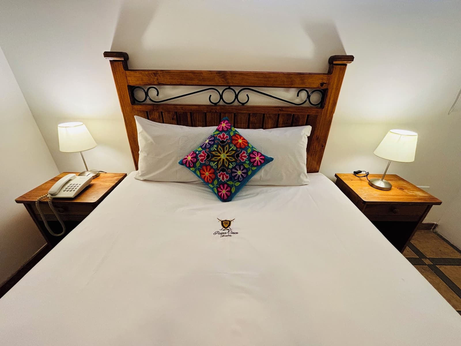 Cama de verdade, lençóis limpos e uma almofada bordada à mão. Merecido. Cama de casal com cabeceira de madeira e ferro forjado, roupa de cama branca, uma almofada andina bordada à mão com cores vibrantes e abajures aconchegantes em um quarto de hotel em Aguas Calientes no trek Salkantay de 5 dias