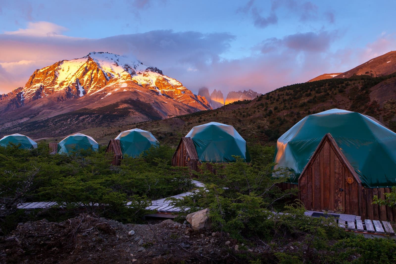 Six domes, one mountain on fire, and the towers deciding whether to show up. A row of teal geodesic domes with rustic wooden entrances at EcoCamp Patagonia, set along a boardwalk amid low shrubs, with snow-capped mountains glowing orange at sunrise and the iconic granite towers of Torres del Paine emerging through clouds in the background — the base for the short w trek.