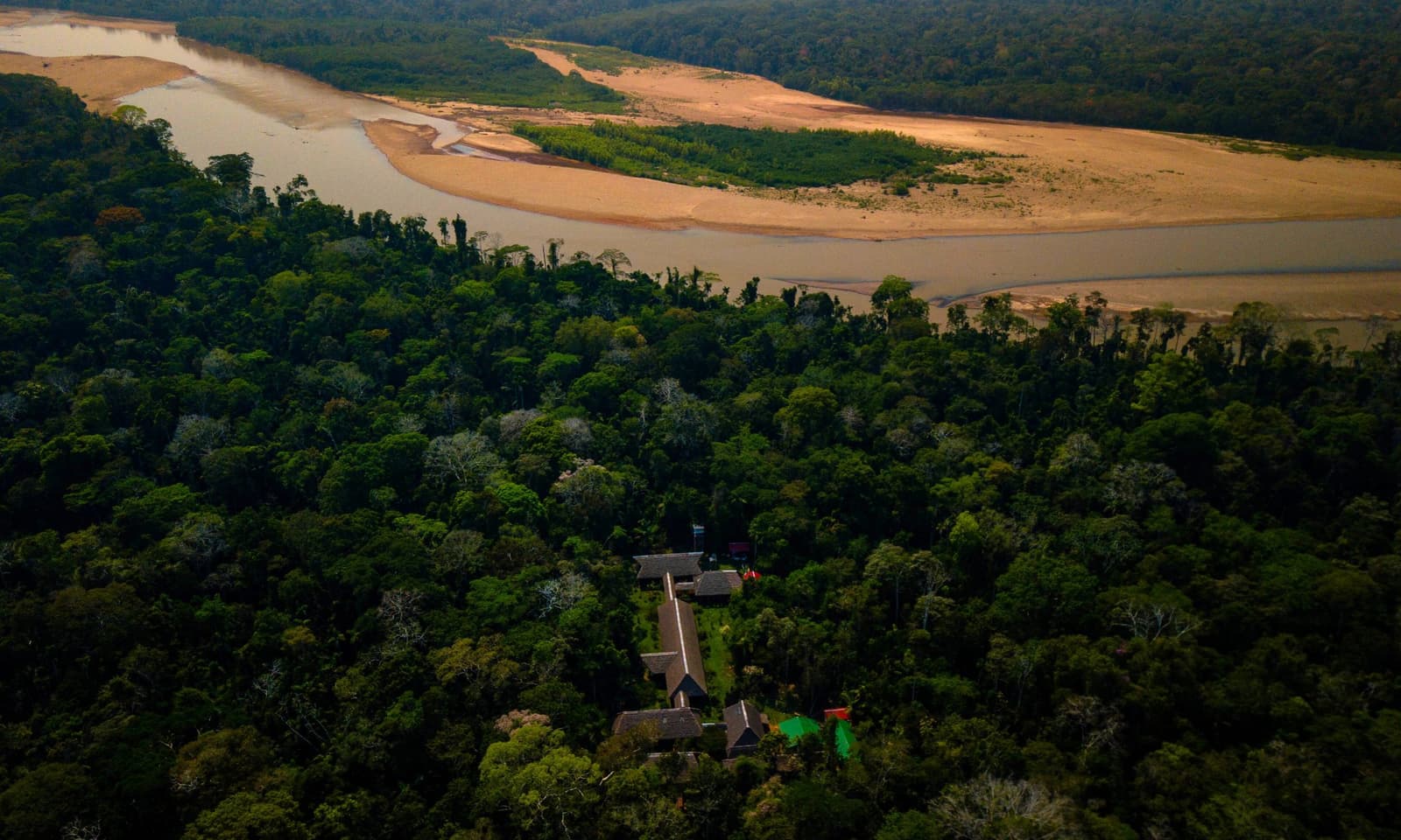 The lodge is in there, small and purposeful, with a whole river bend to itself. Aerial view of the Tambopata Research Center Peru lodge nestled in dense Amazon rainforest canopy beside a wide river with golden sandbanks curving through the jungle