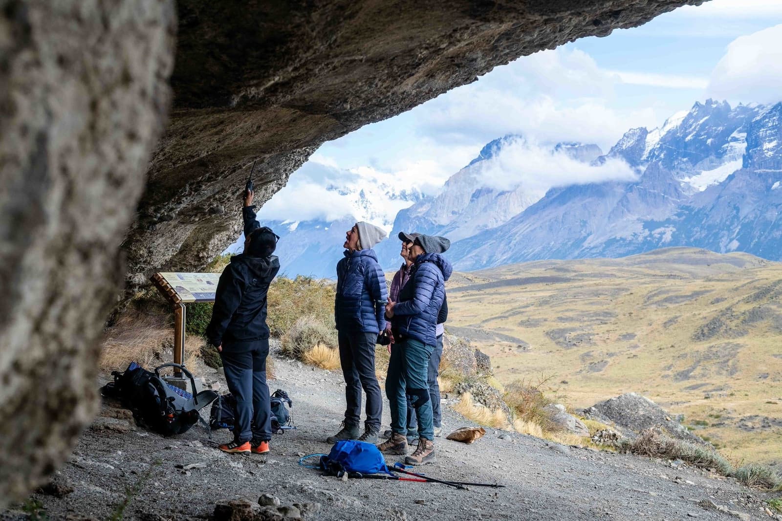 A cave with a view, your guide knows every story the rock has to tell. A guide points a torch at the rock ceiling of a cave shelter while three guests look up, with the snow-capped peaks of Torres del Paine visible across the open steppe behind them on a torres del paine 4 day all inclusive tour