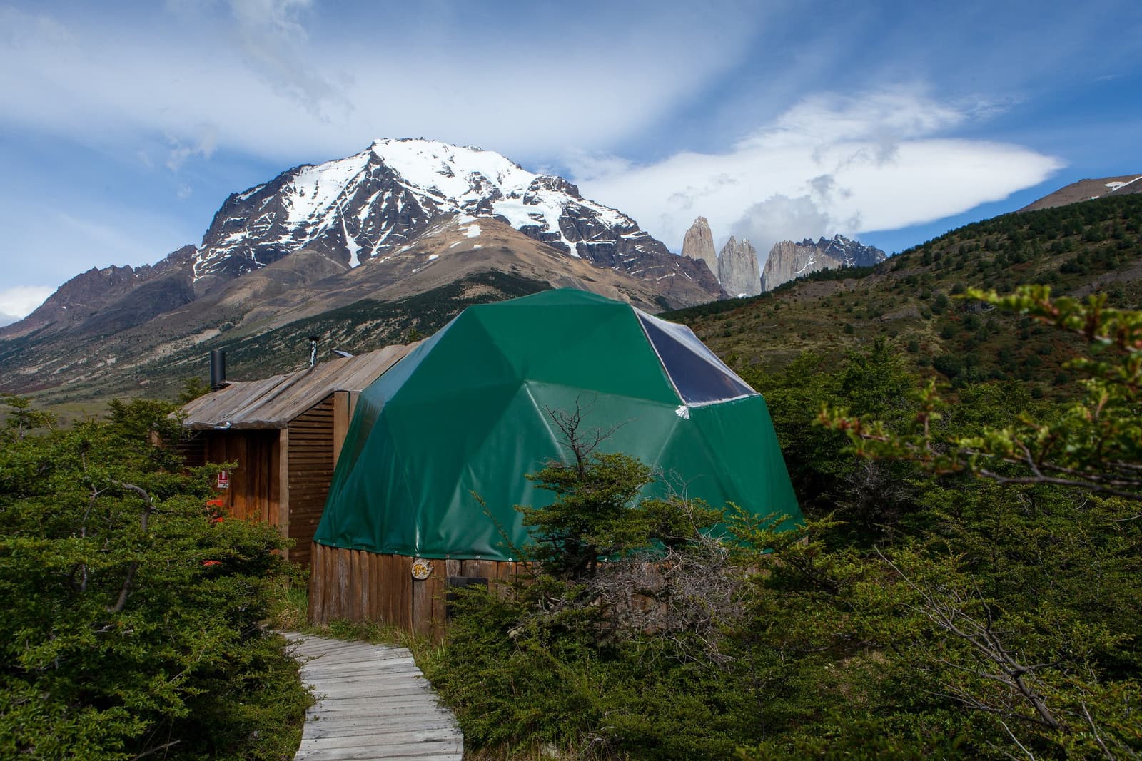 Your room has a better backdrop than most screensavers. Photo: Superior Dome A green geodesic dome with a wooden base sits along a boardwalk path surrounded by Patagonian shrubs, with a snow-capped mountain and the three granite towers of Torres del Paine visible in the background, part of this torres del paine 4 day all inclusive tour at EcoCamp Patagonia