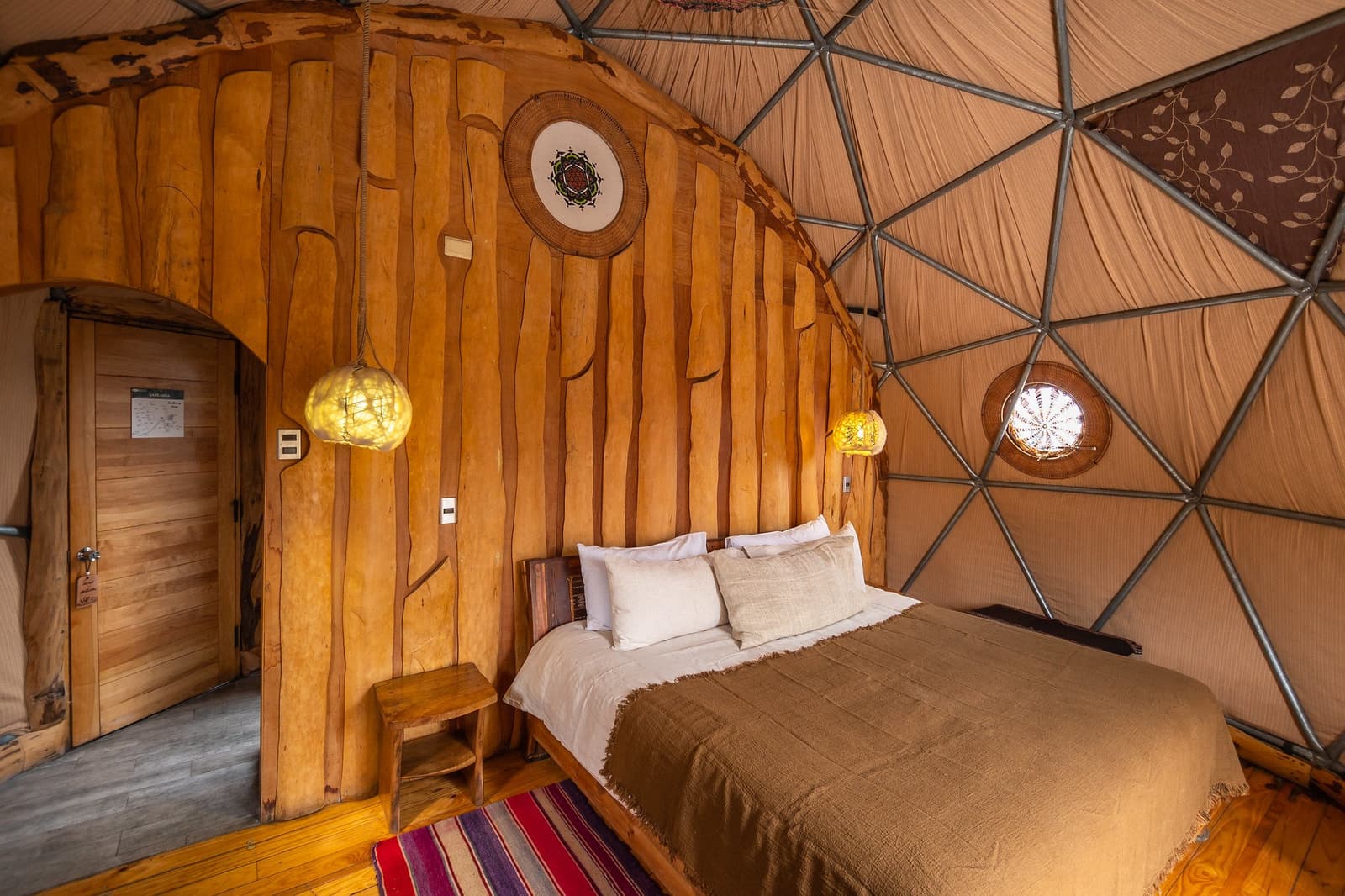 Handcarved wood, warm light, and a bed that earns its keep after a long day on the trail. Photo: Suite Dome Interior of a geodesic eco-dome on a torres del paine 4 day all inclusive tour, featuring a double bed with linen pillows, handcrafted wooden wall panels with organic carvings, warm woven pendant lights, a colorful striped rug, and the dome's triangular metal-and-canvas ceiling structure