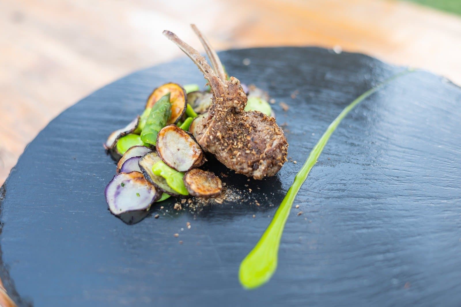 Patagonian lamb, done properly, on a slate board that means business. Herb-crusted lamb cutlets plated on a slate board with roasted purple potato rounds, green vegetables, and a vivid green sauce swipe, part of the torres del paine 4 day all inclusive tour dining experience at EcoCamp Patagonia