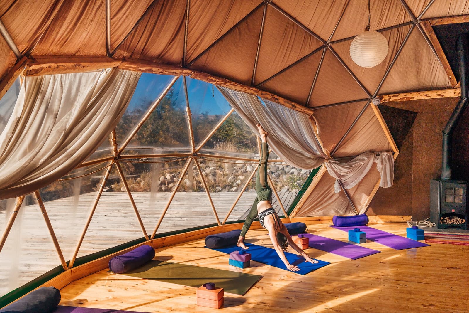 O sol entra pelos painéis do domo toda manhã, e o tapete te espera. Photo: Yoga Dome Uma mulher pratica a postura de cachorro olhando para baixo em um tapete azul dentro de um domo geodésico com piso de madeira, cortinas de linho, blocos de yoga e vista para a vegetação patagônica durante o trekking W de Torres del Paine