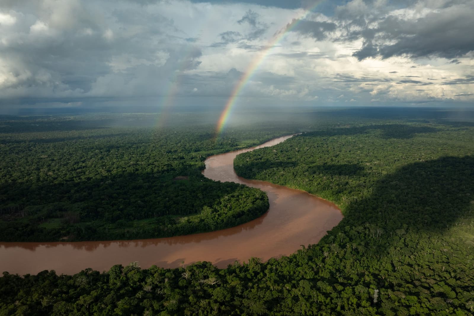 Rain just passed, the river doesn't care, and the forest carries on. Aerial view of a wide, sediment-rich river winding through dense rainforest canopy on an amazon tour from Puerto Maldonado, with a vivid rainbow arching over the horizon beneath a dramatic storm sky