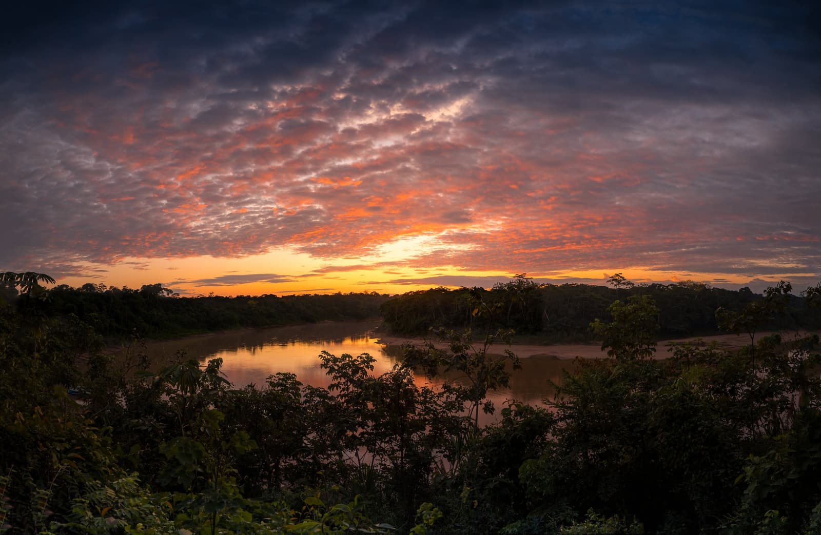 The river keeps the score at the end of every good day. Photo: GABRIEL_S Wide panoramic view of a winding jungle river reflecting an orange and red sunset sky, surrounded by dense rainforest canopy on an amazon tour from Puerto Maldonado