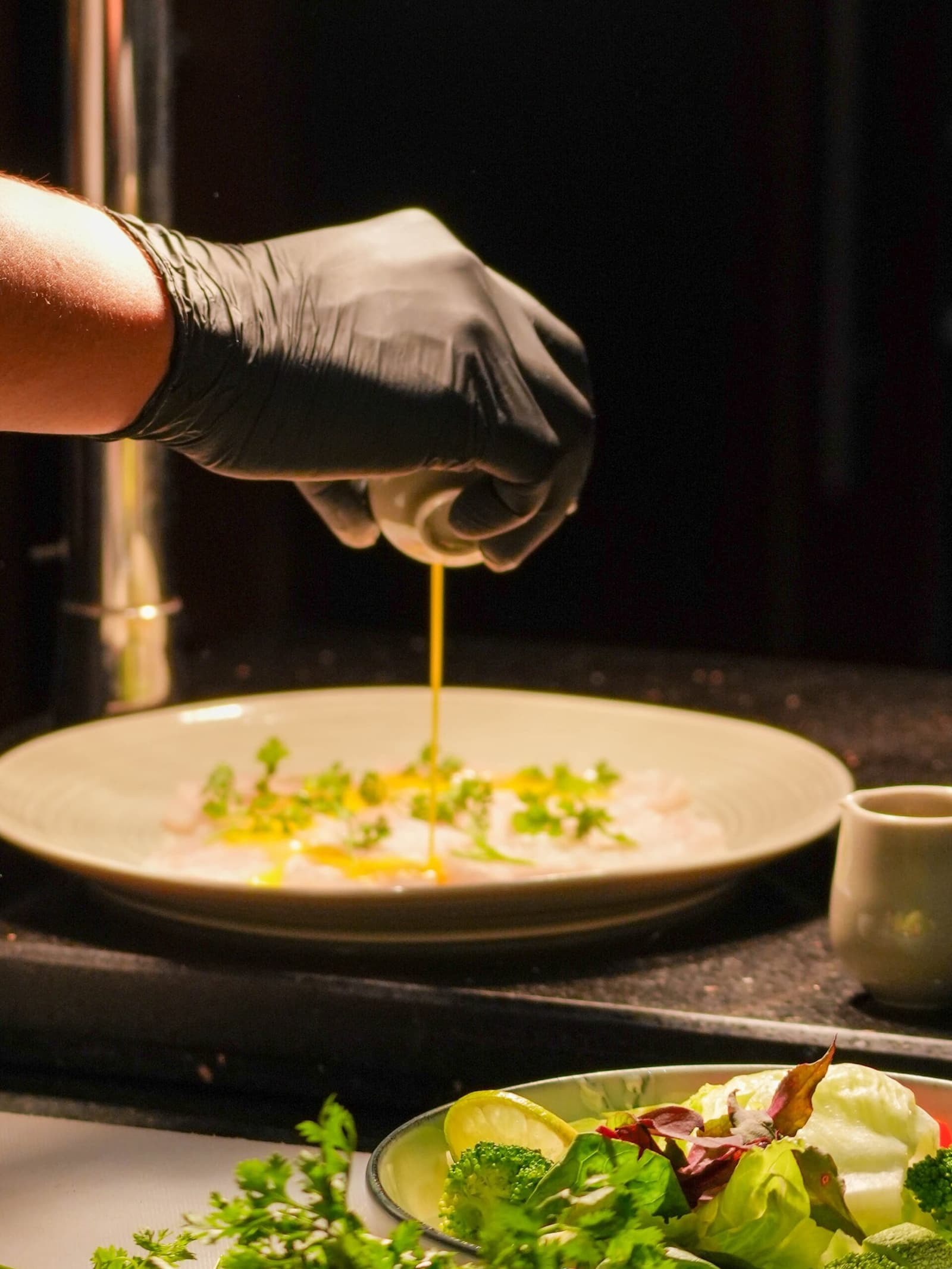 Good ingredients, a steady hand, and the river outside the window. Photo: Aqua Expeditions A chef's gloved hand drizzles golden oil over a delicate fish carpaccio garnished with fresh herbs, plated on the Aria Amazon cruise, with a side salad and small sauce cup nearby