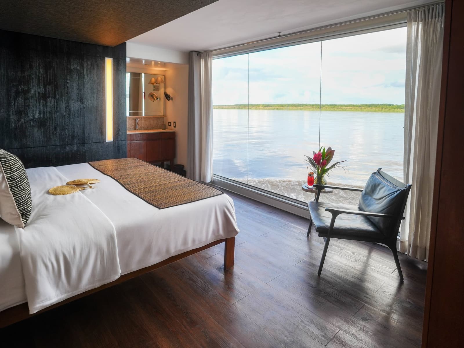 Waking up to that view is not a feature, it is the whole point. Photo: Francois Moreau Spacious cabin on the Aria Amazon cruise with a king bed, hardwood floors, and a floor-to-ceiling glass wall opening directly onto a wide, calm river bordered by low jungle vegetation