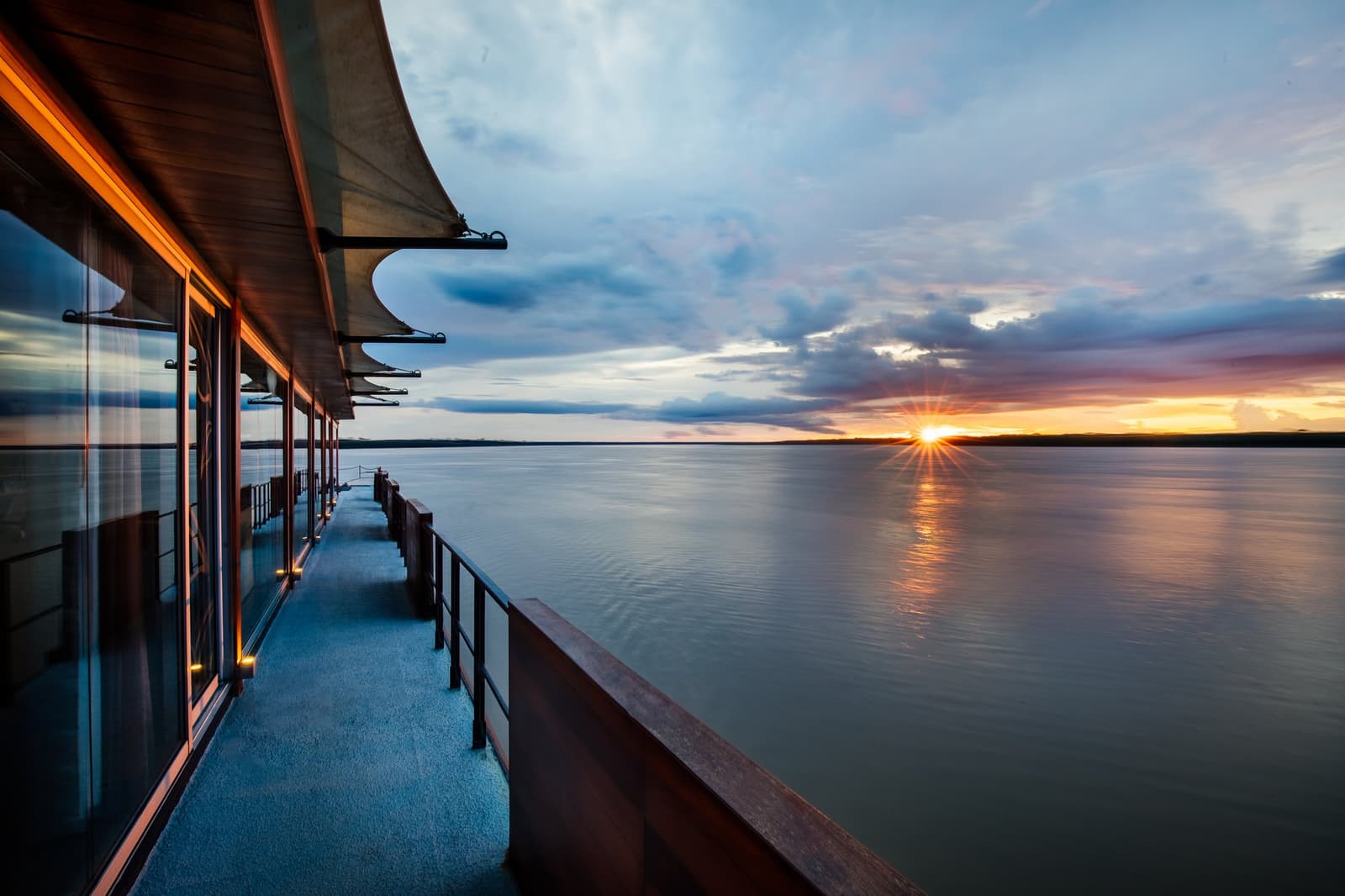 The river gets the last word every evening, and the deck puts you front row. Photo: Aqua Expeditions The open promenade deck of the Aria Amazon cruise ship stretches toward the bow, with floor-to-ceiling cabin windows to the left and a wide river glowing under a vivid sunset on the horizon