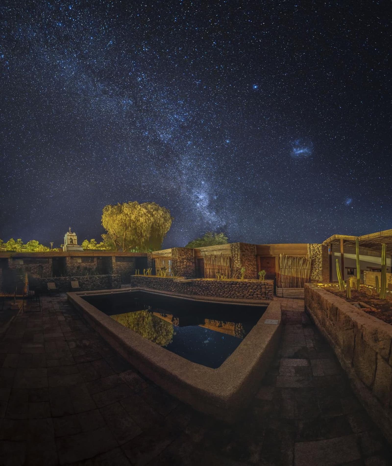 La piscina refleja la galaxia. El desierto tiene sus propias recompensas. Patio de un lodge de piedra y caña con una piscina oscura que refleja el cielo nocturno, la Vía Láctea arqueándose sobre las estrellas encima de una base de camping en el desierto de Atacama, con una torre de iglesia iluminada y árboles brillantes al fondo