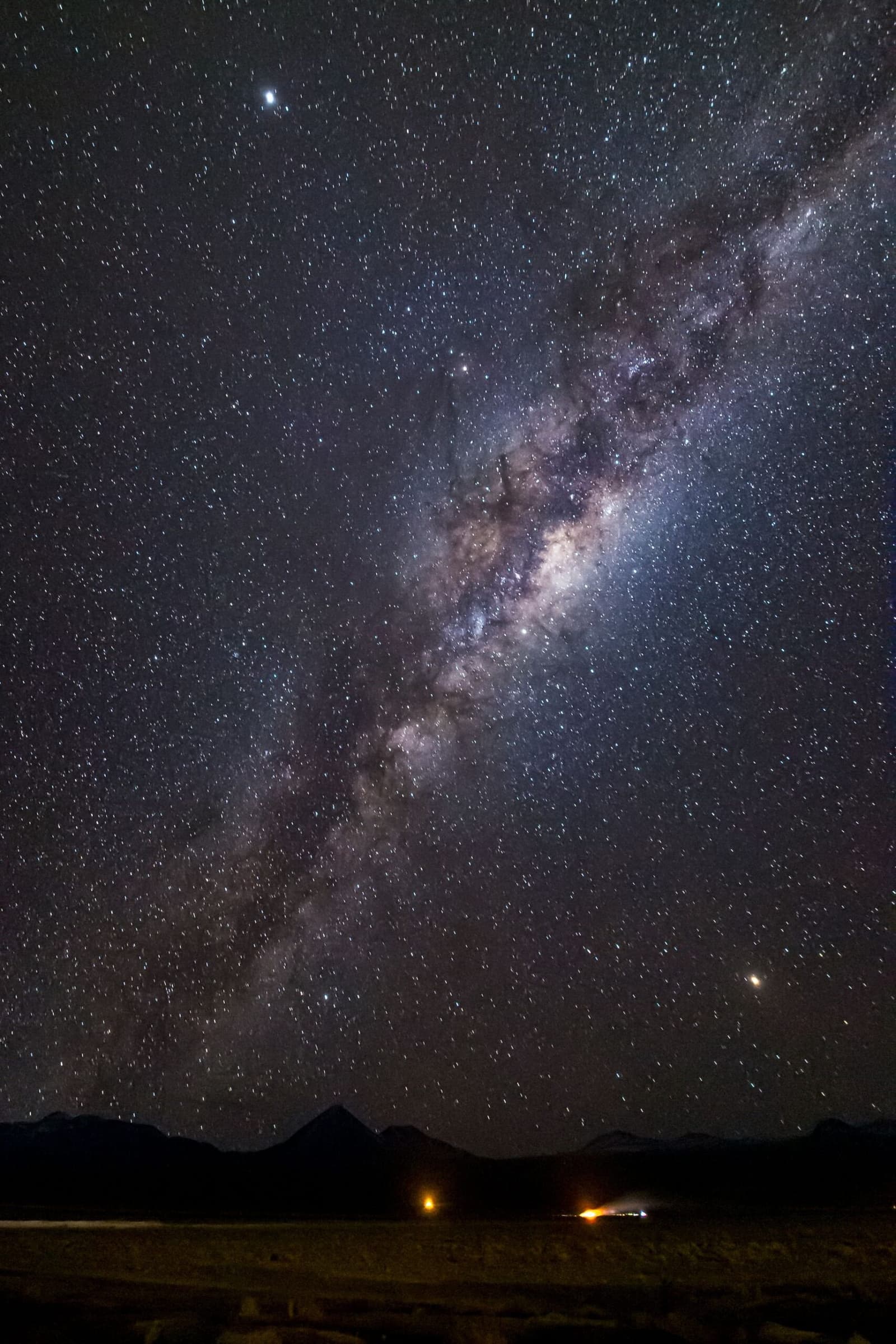 Sin contaminación lumínica, sin vecinos, exactamente para esto viniste. Photo: Erike Fusiki El núcleo galáctico de la Vía Láctea forma un arco sobre la silueta oscura de las montañas durante el camping en el desierto de Atacama, con dos puntos de luz cálida visibles en la llanura distante