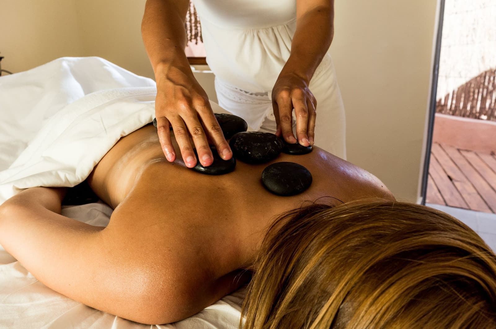 The desert works you hard. This is the part where you stop and let someone else do the work. A therapist places warm black basalt stones along a guest's back during a hot stone massage at a lodge spa, with a sunlit wooden deck visible through an open door in the background — a relaxing add-on to an atacama desert tour package
