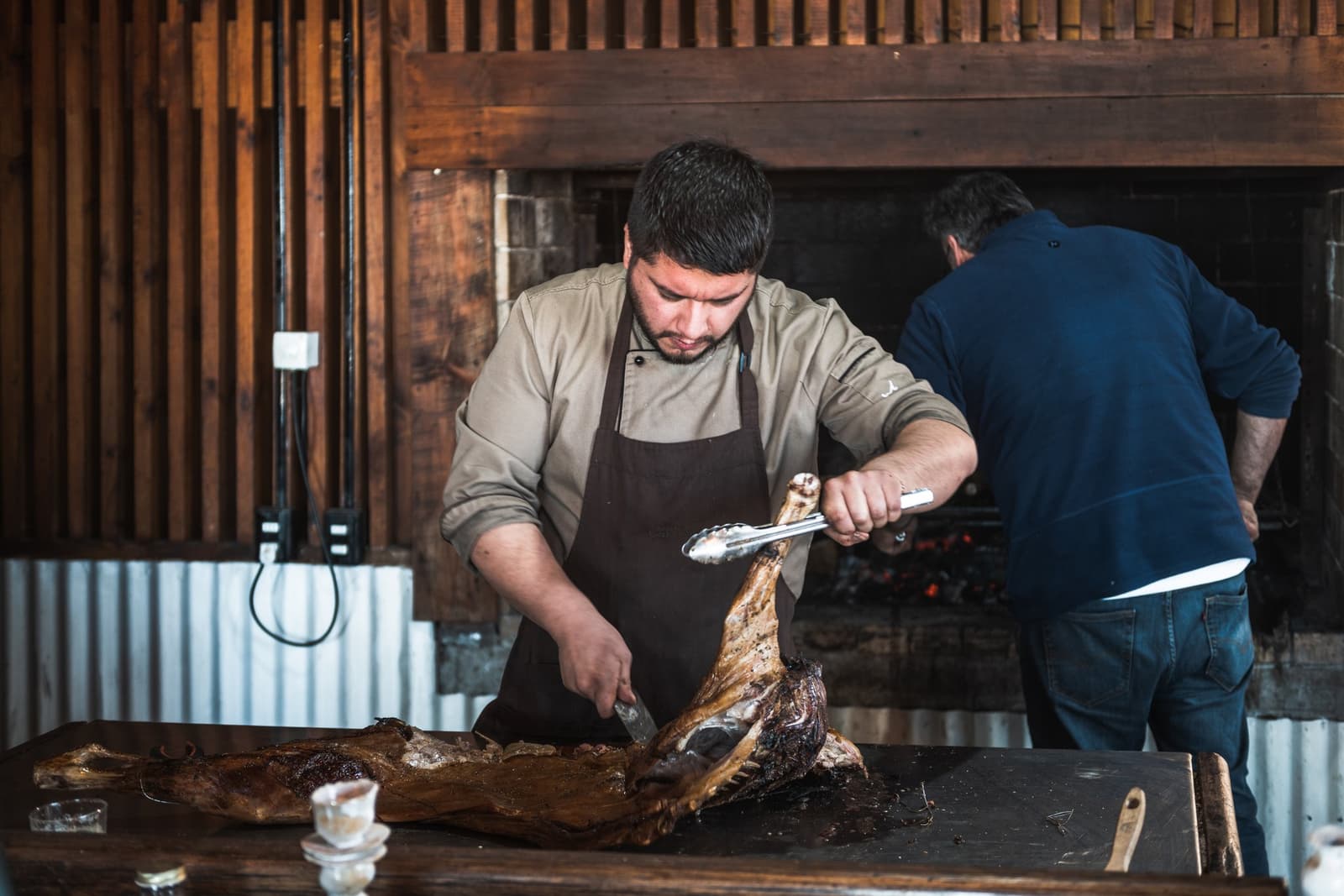 Whole lamb, open fire, a knife doing the talking. Photo: CARLOS DIAZ A chef in a brown apron carves a whole fire-roasted lamb at a wooden table inside the estancia cerro guido dining room, with glowing embers visible in the stone fireplace behind him