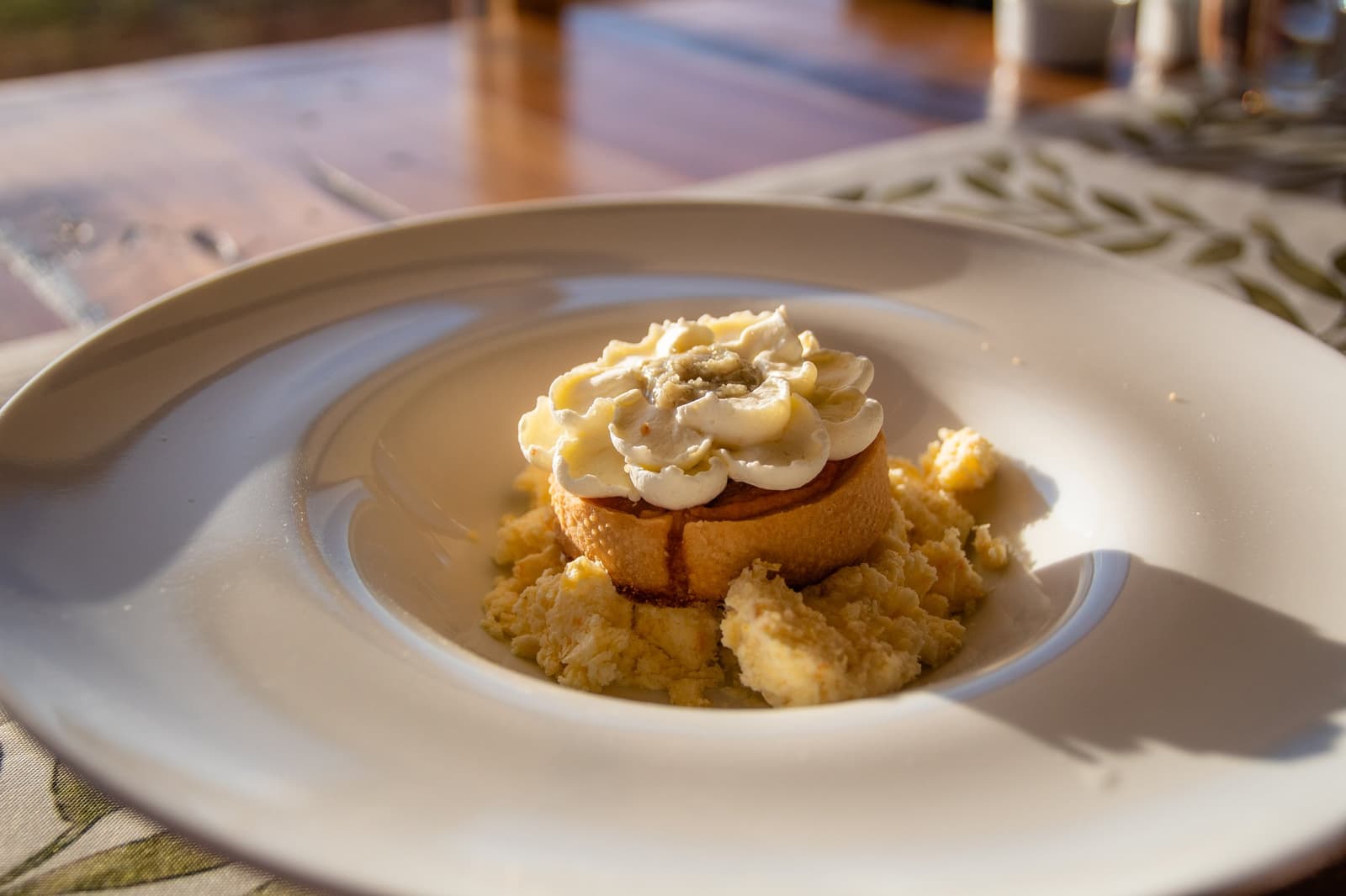 Dessert that earns the long drive from Puerto Natales. A golden sponge cake round topped with piped cream and a crumble base, plated on a wide white dish at Estancia Cerro Guido, lit by warm afternoon light